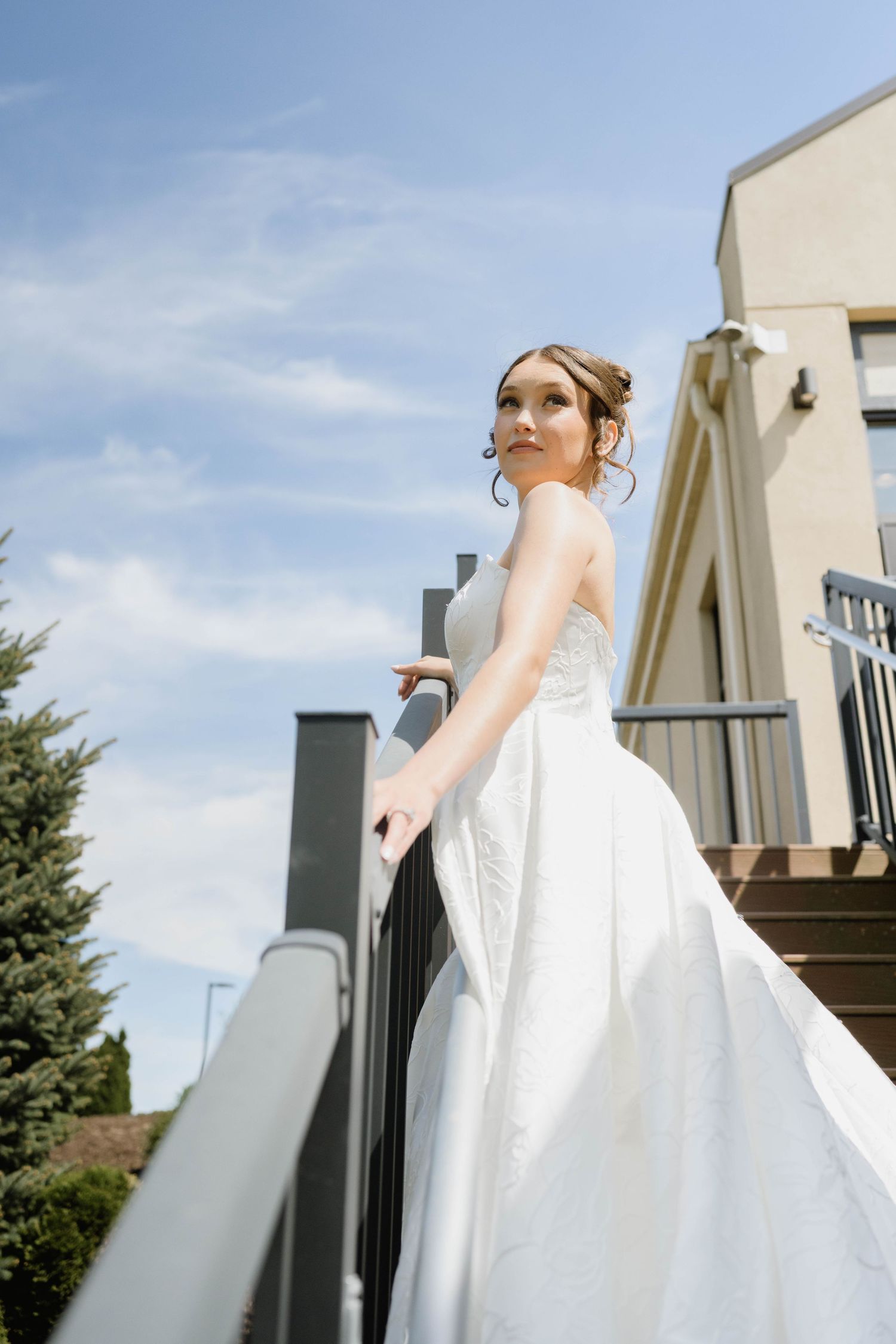 Beautiful bride in white ball gown standing on outdoor stairs against a blue sky.