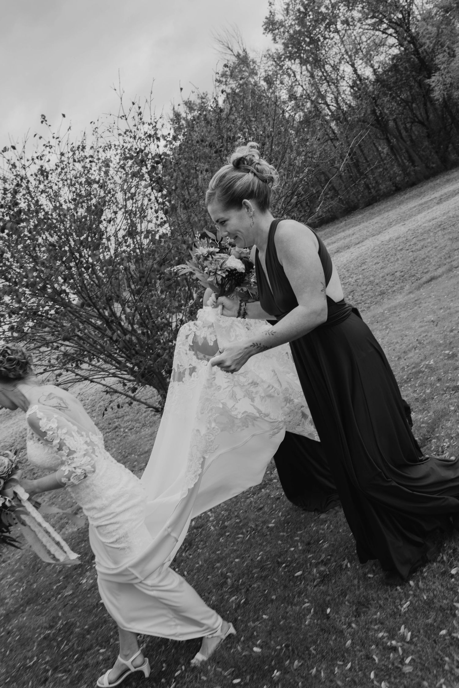 Bridesmaid in a dark dress holds a bouquet and helps lift the bride's lace wedding gown train outdoors on a windy day in black and white.