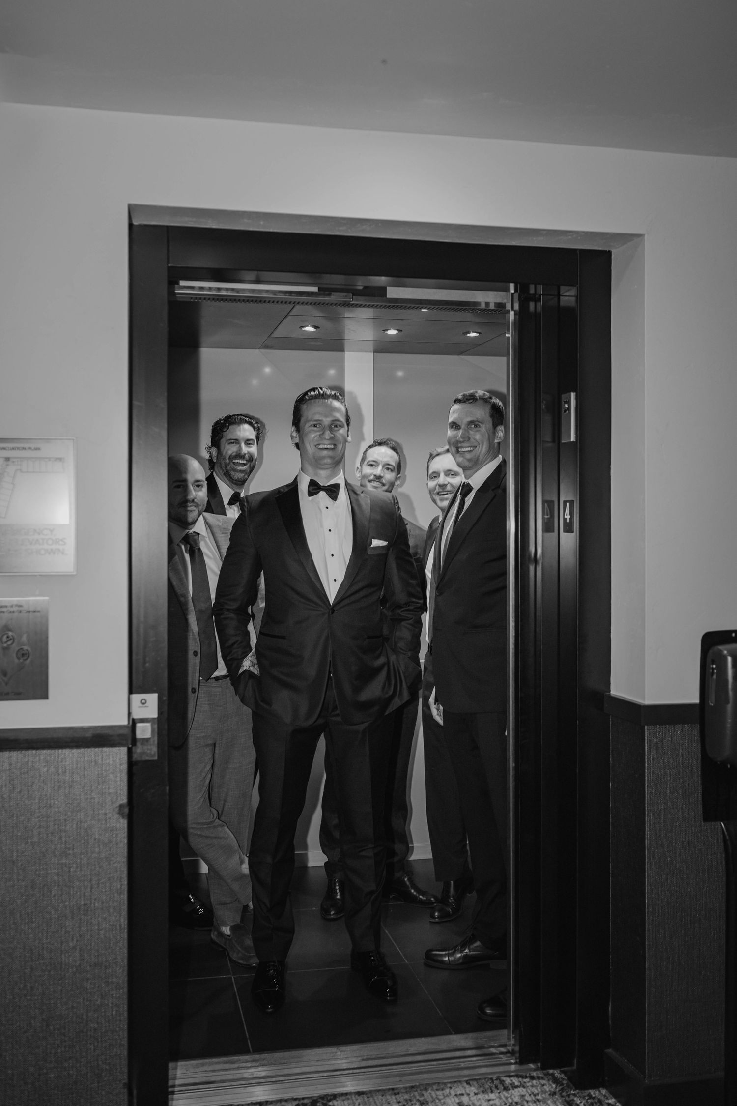 Black and white photo of a groom in a tuxedo exiting an elevator with groomsmen behind him.