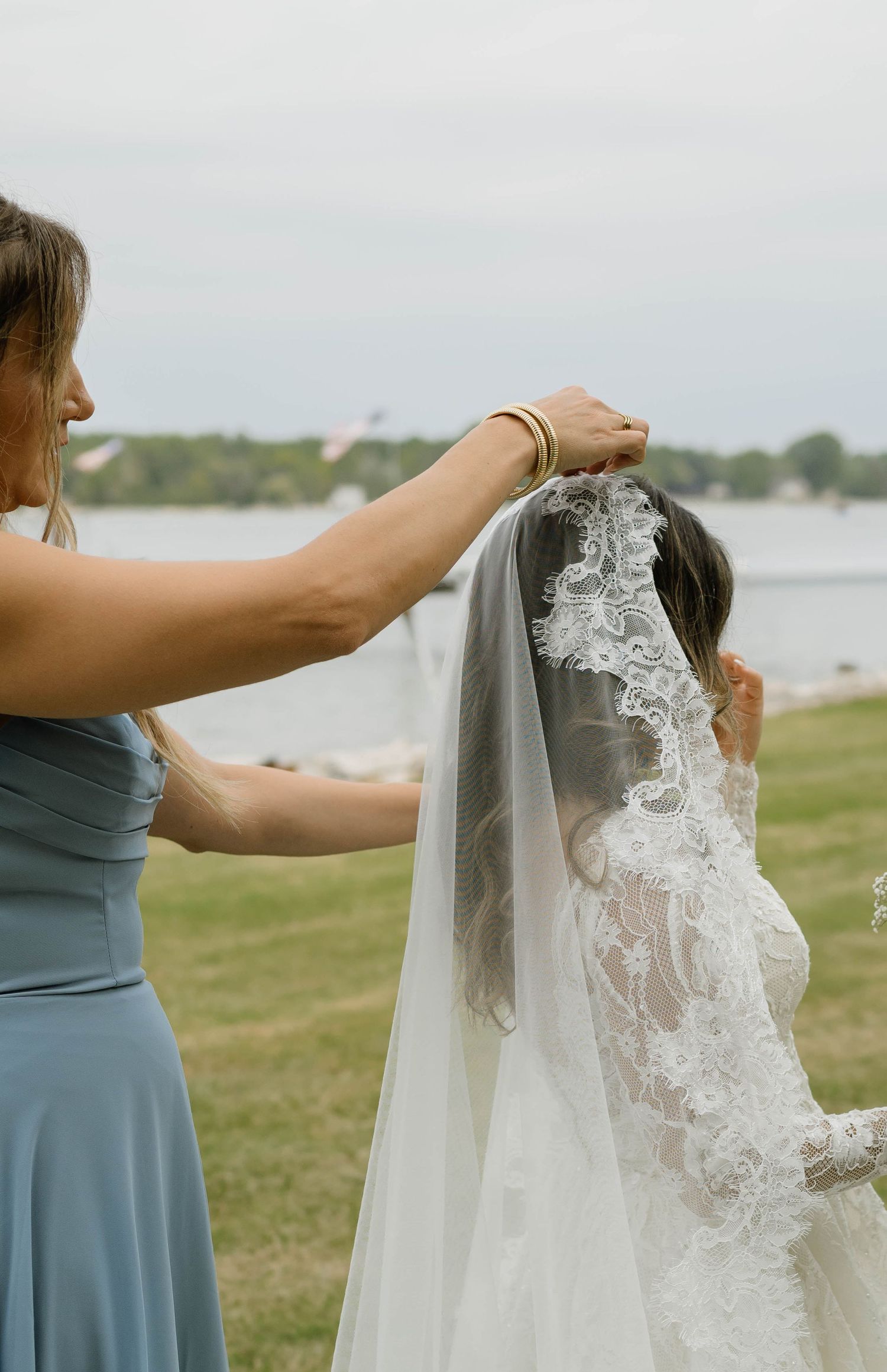 Bridesmaid adjusting lace cathedral wedding veil on bride outdoors near a waterfront venue.