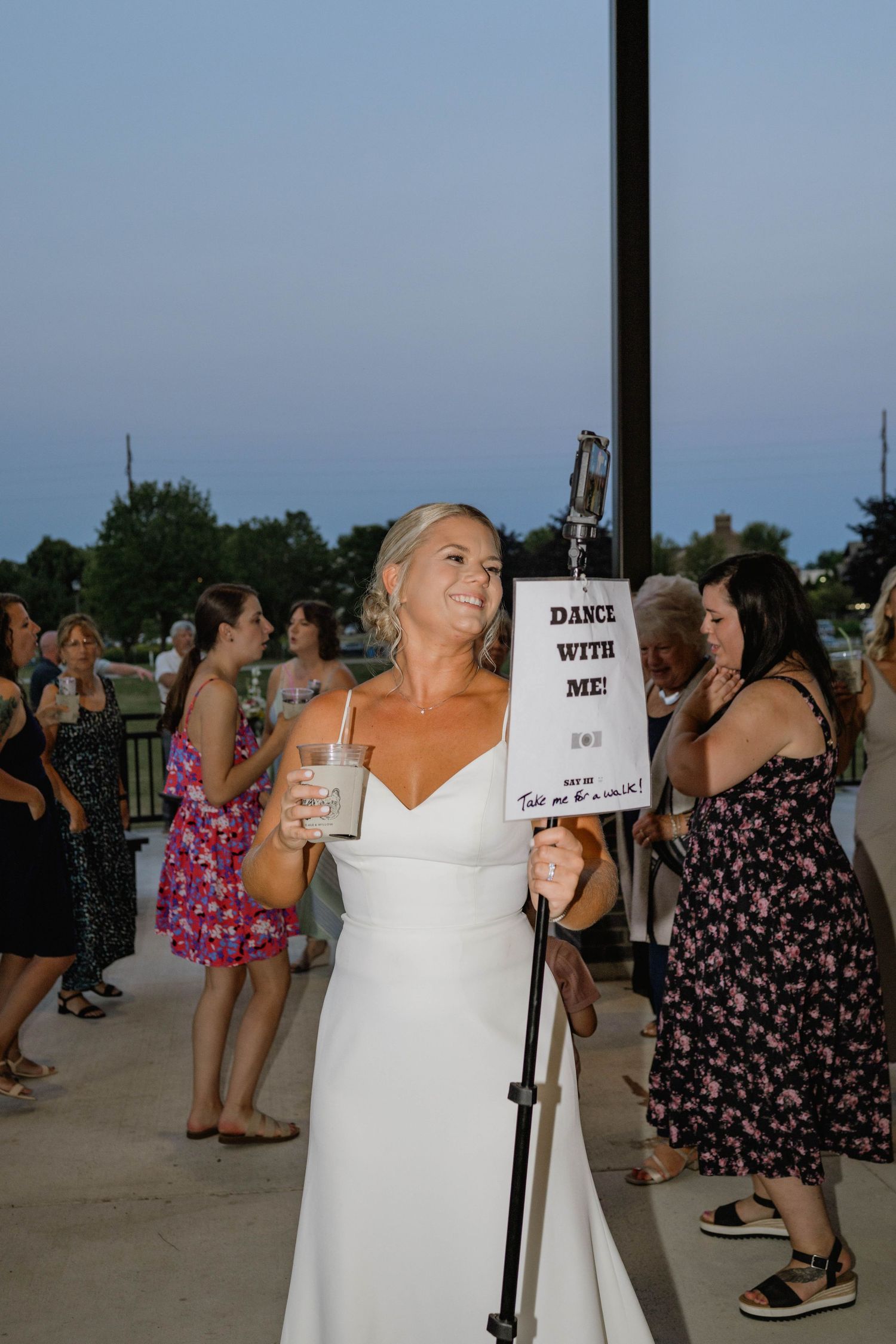 Smiling bride in white gown holds drink and dance invitation sign at outdoor wedding reception.