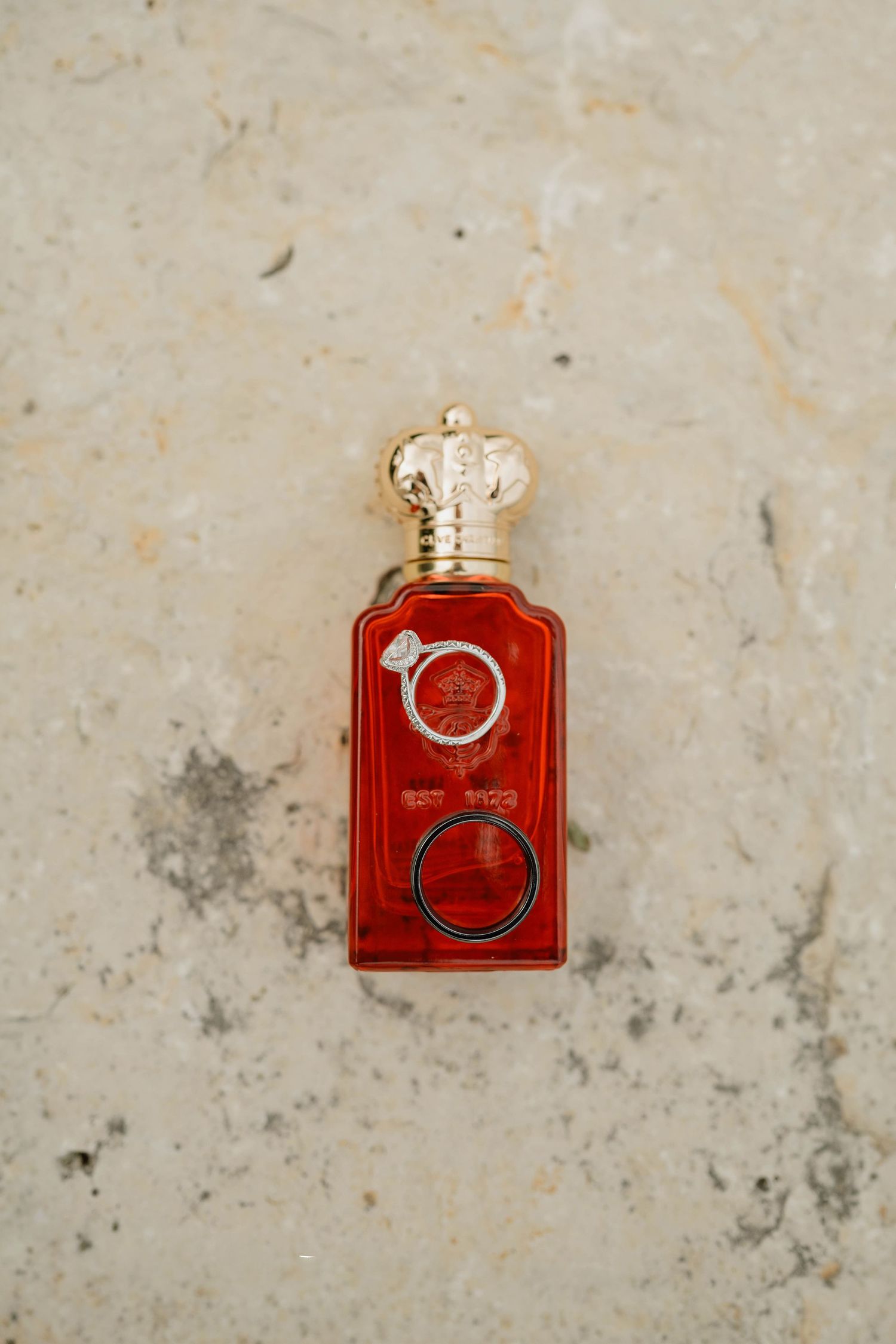 Two wedding rings rest atop a red and gold ornate perfume bottle on marble.