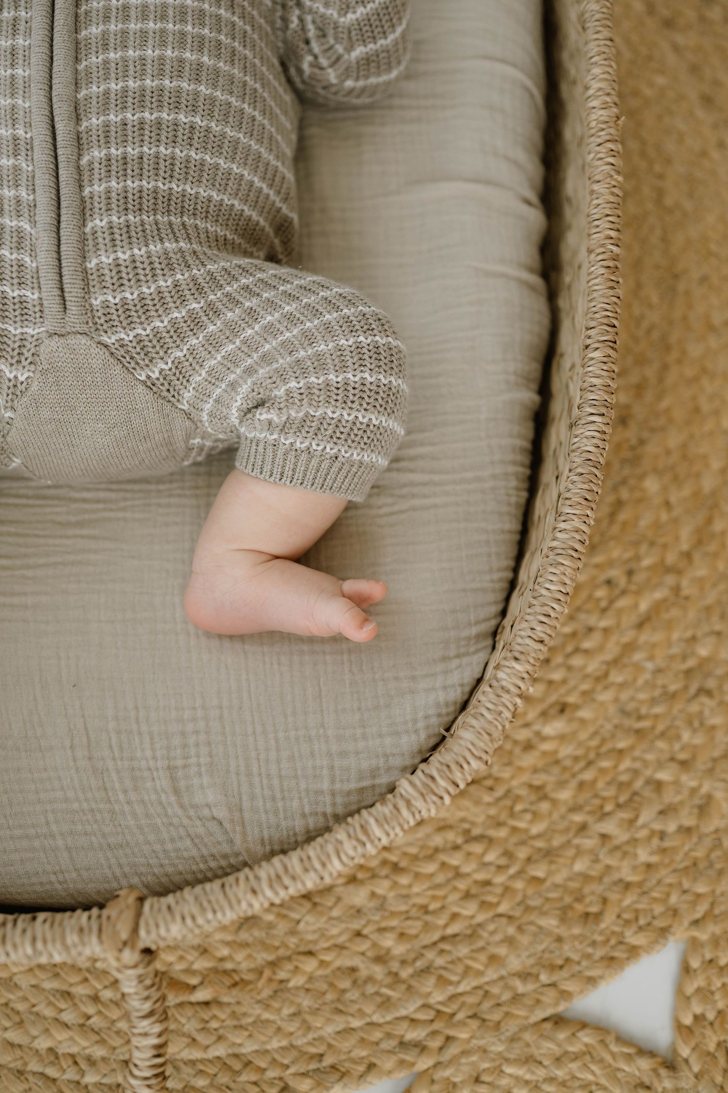 Sleeping newborn baby in striped knit onesie resting in a woven bassinet with sage green lining.