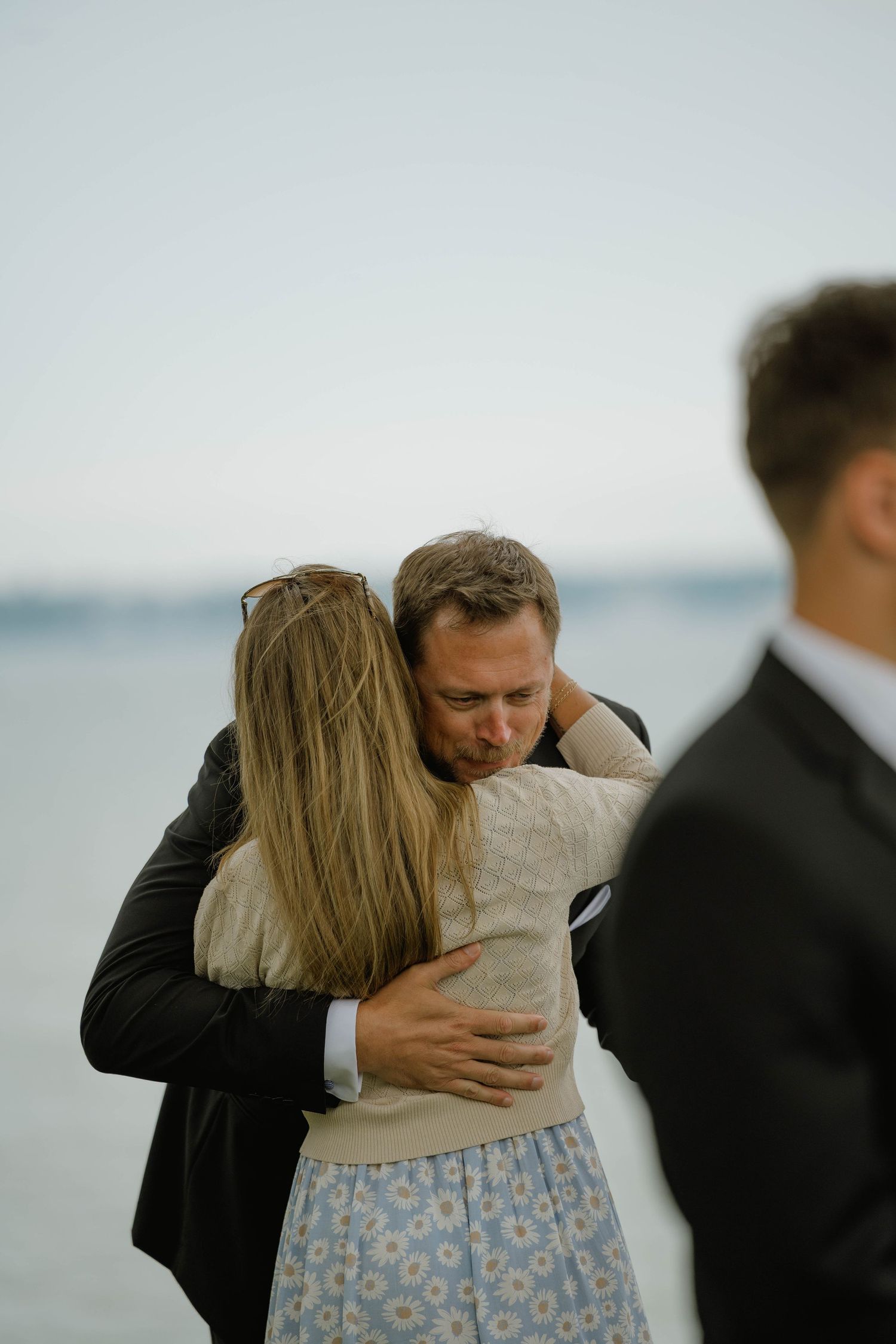 Emotional wedding moment as groom embraces a female guest near the waterfront ceremony.