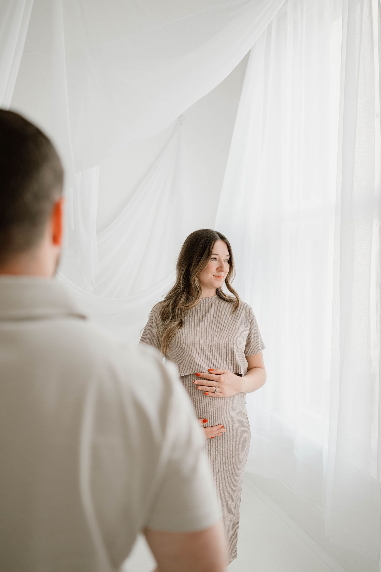 A pregnant woman in a beige dress cradles her bump while her partner looks on.