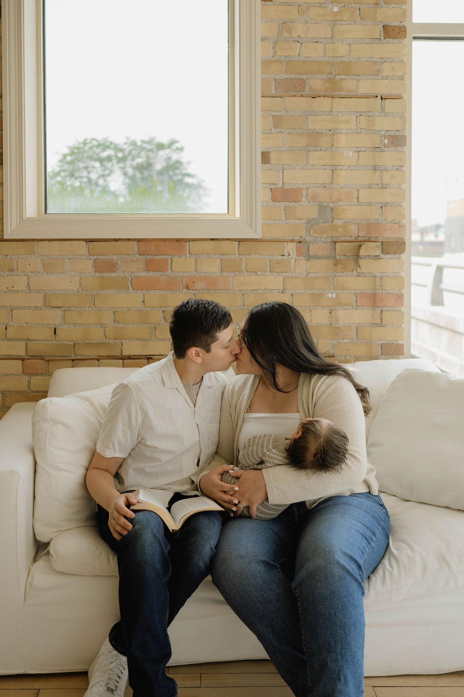 A couple shares a tender kiss on a white couch while cradling their newborn baby in a cozy brick-walled living room.