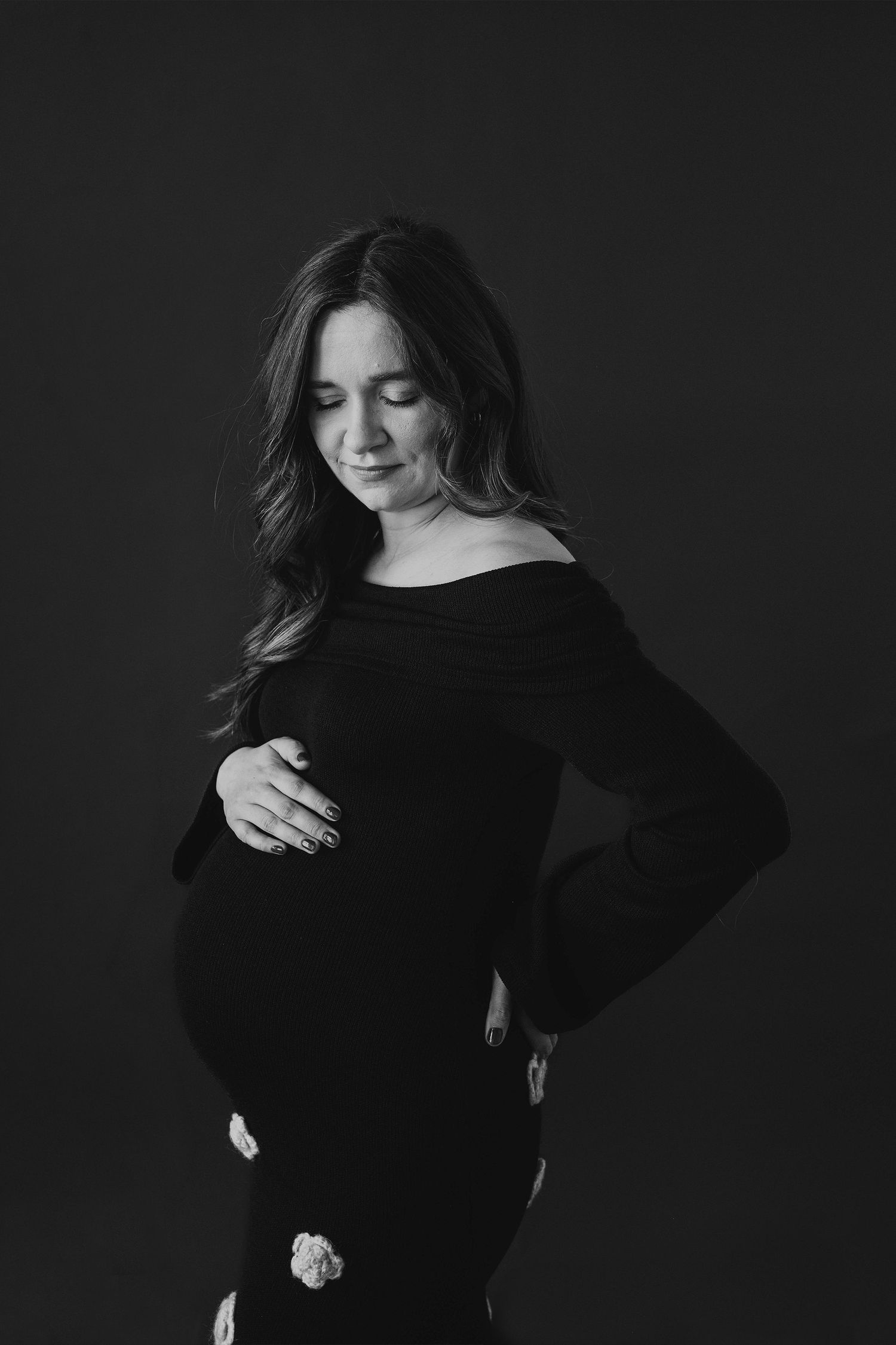 Black and white maternity portrait of a pregnant woman in an off-shoulder dress gently cradling her baby bump against a dark backdrop.