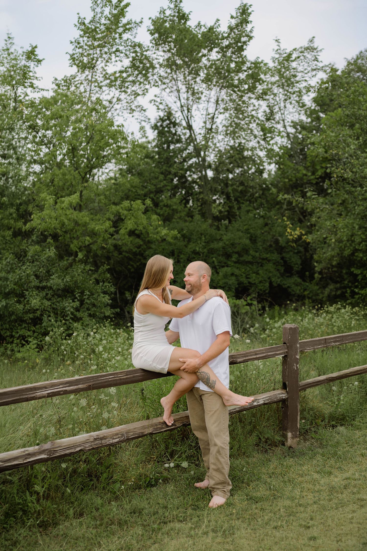 Couple sharing romantic moment sitting on rustic wooden fence surrounded by lush greenery.
