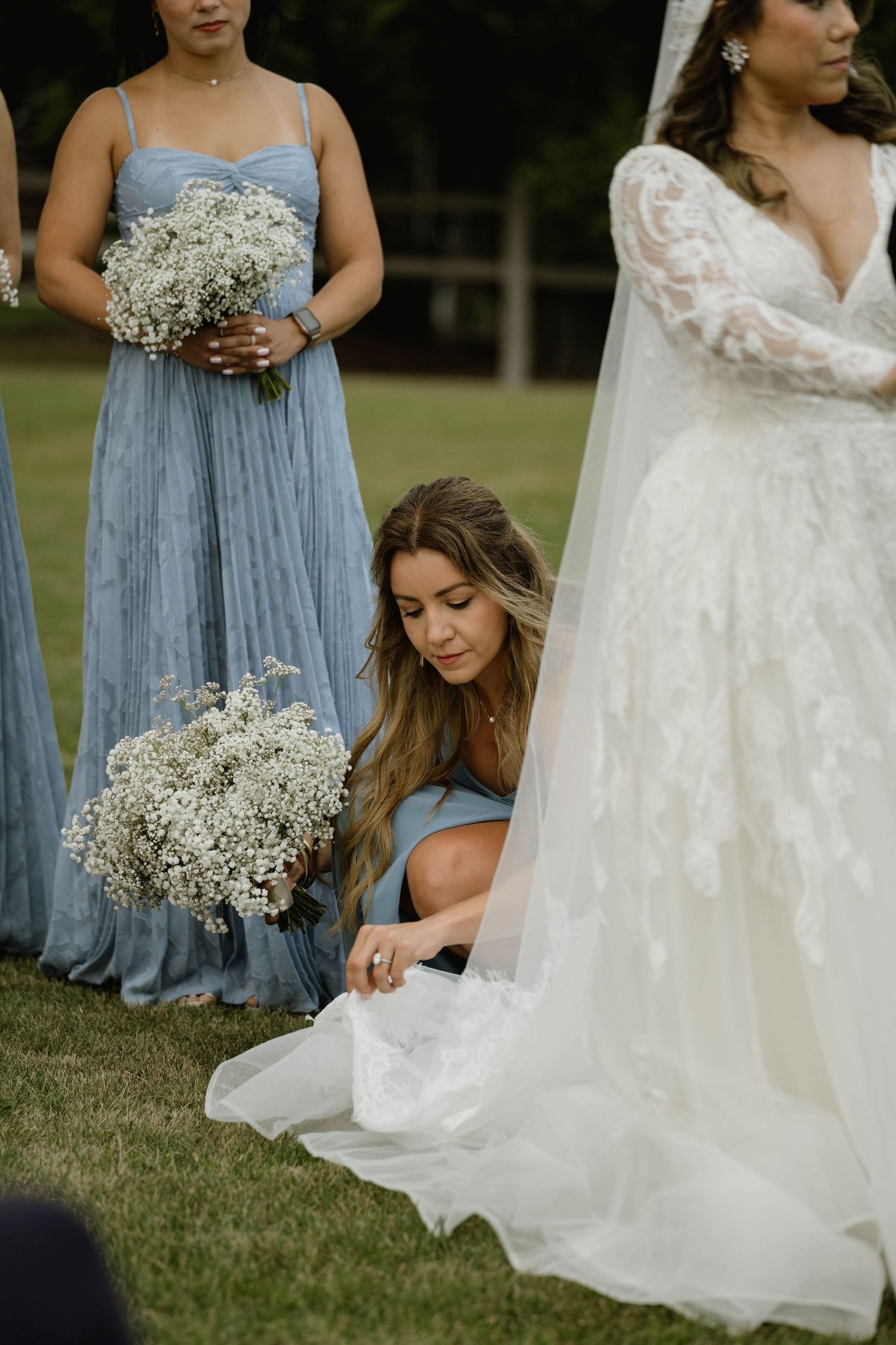 Bridesmaid in blue dress adjusts bride's white lace wedding gown train on grass at outdoor ceremony.