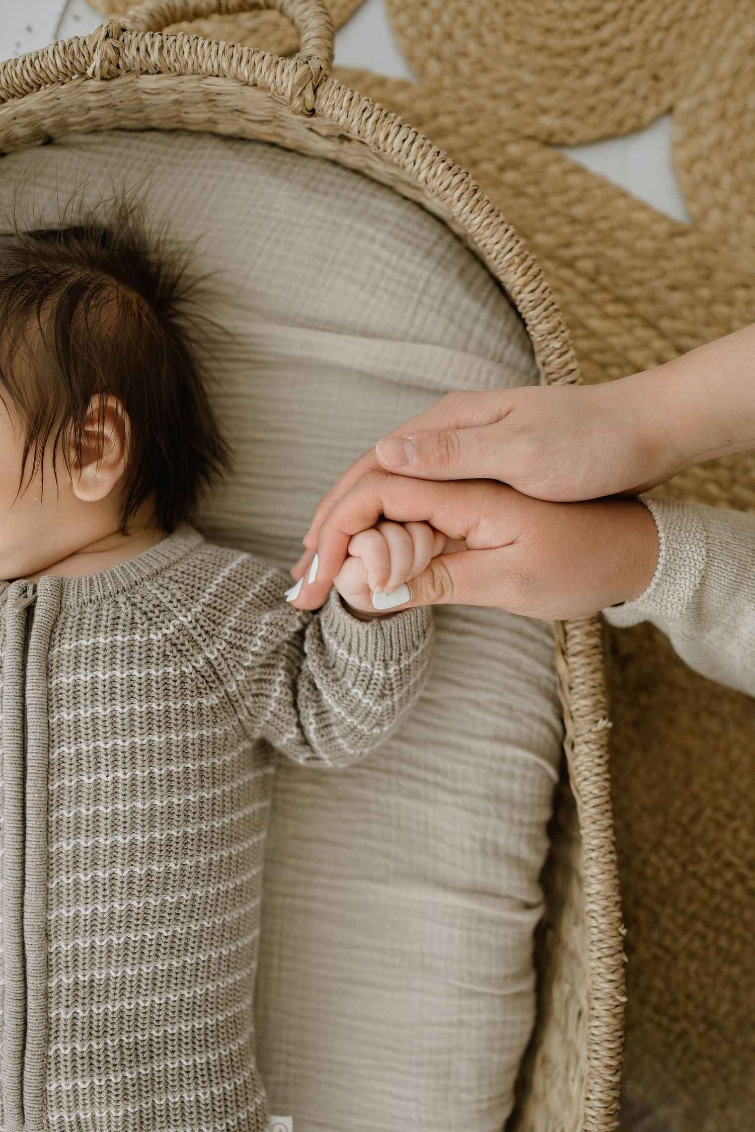 Parent gently holds sleeping newborns tiny hand while baby rests in a woven bassinet.