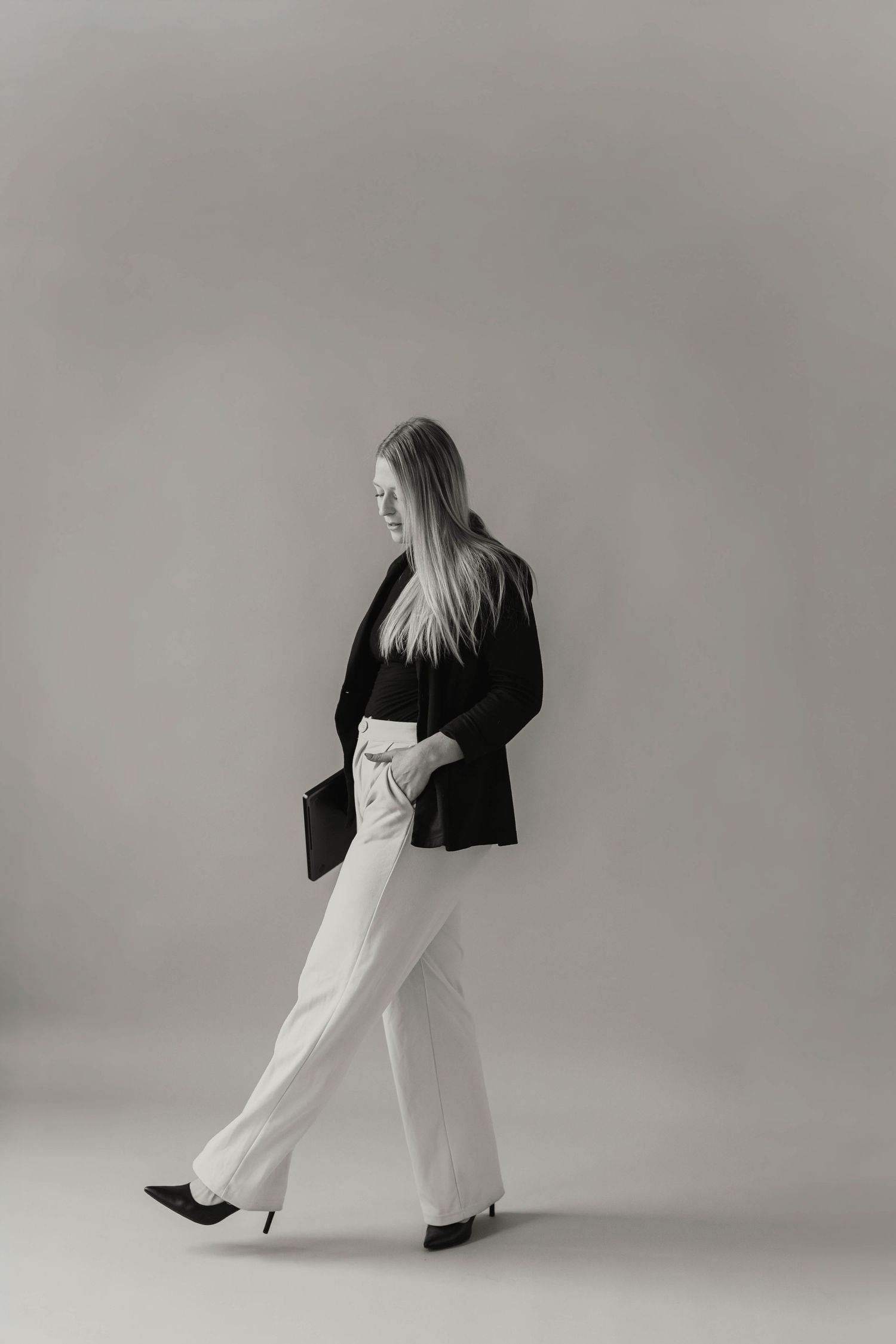 Black and white side profile of a blonde woman walking in white wide-leg pants and black blazer.