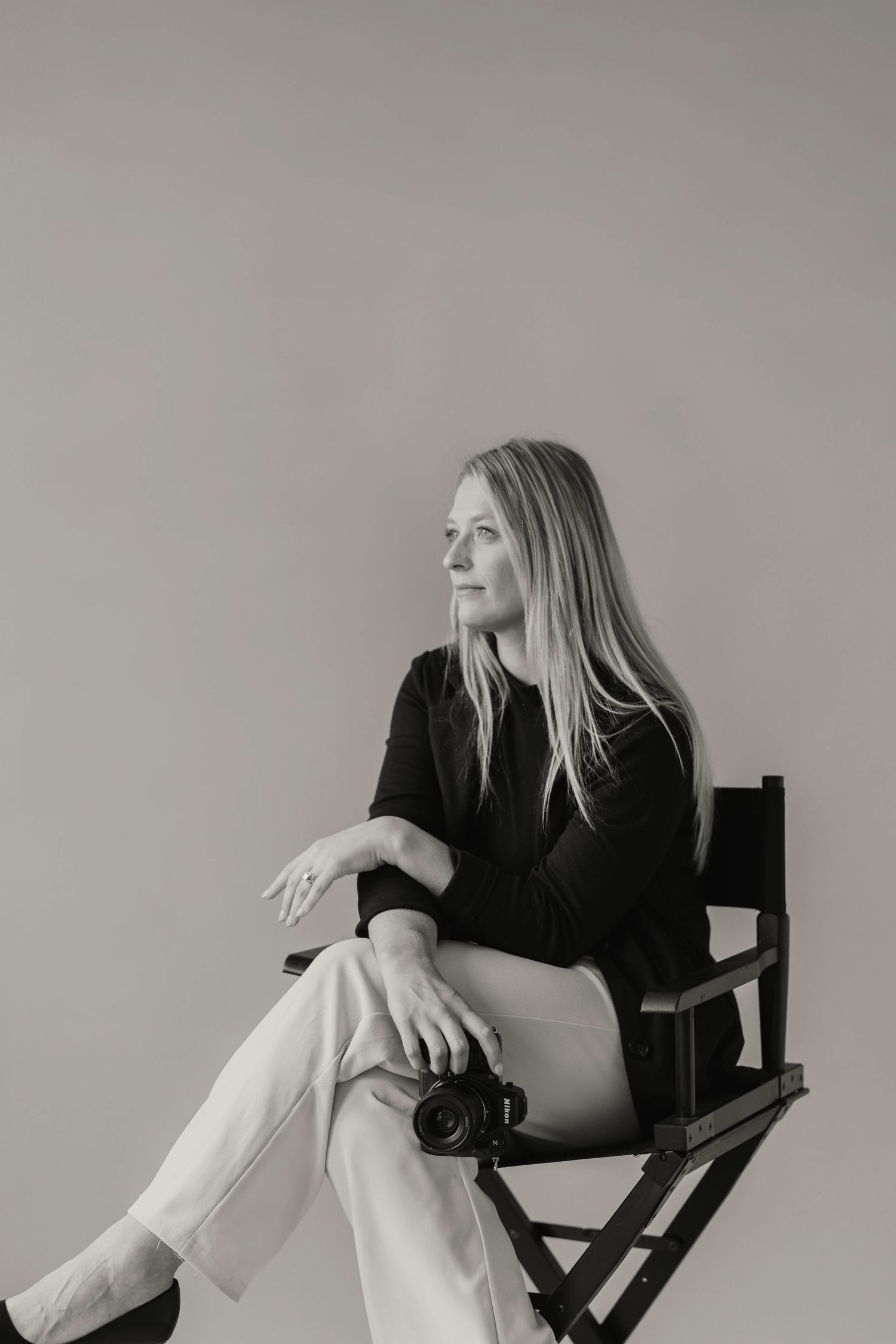Black and white portrait of a blonde female photographer sitting in a director's chair with camera.