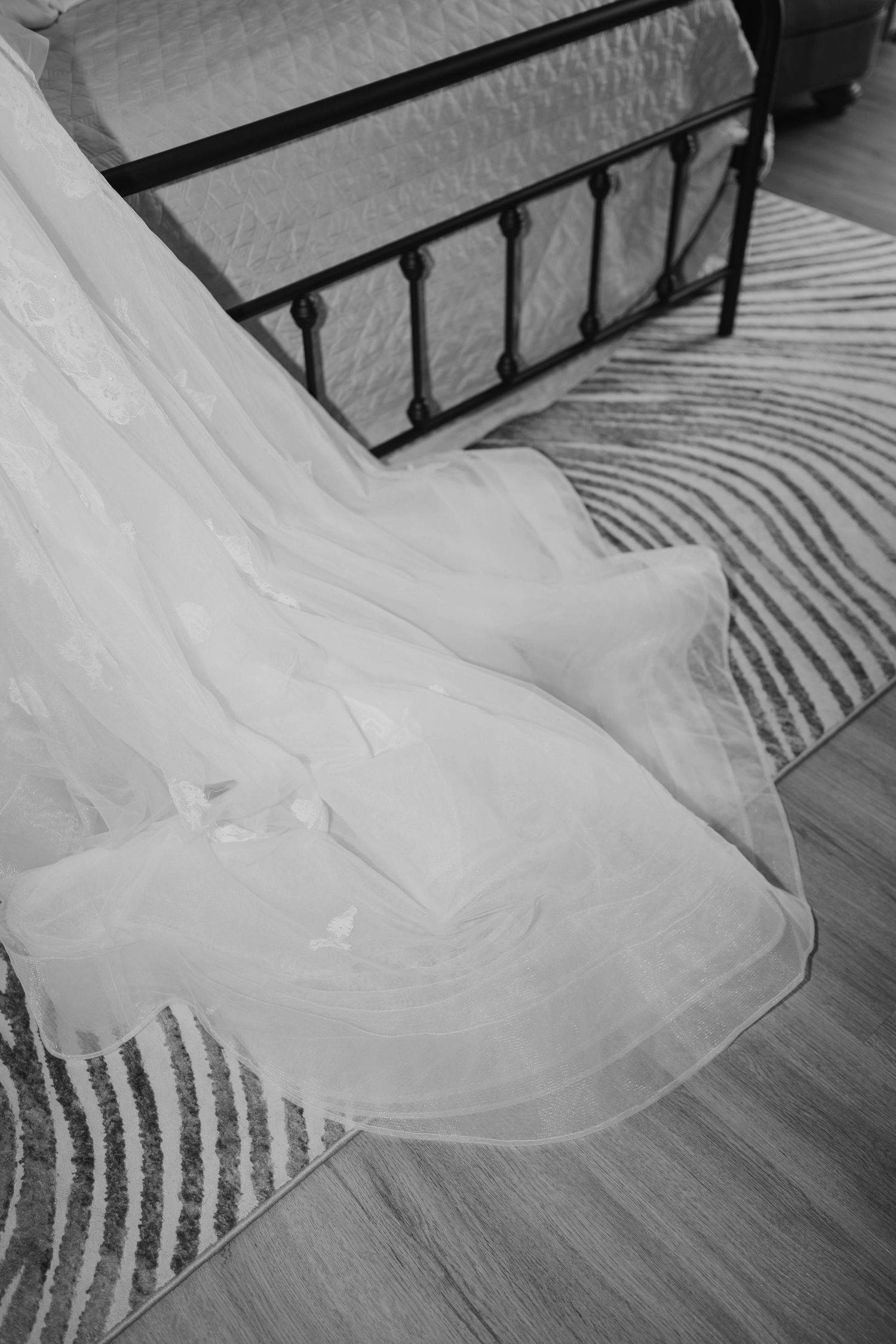 Black and white close-up of a delicate white wedding veil draped across a striped rug beside a black iron bed.