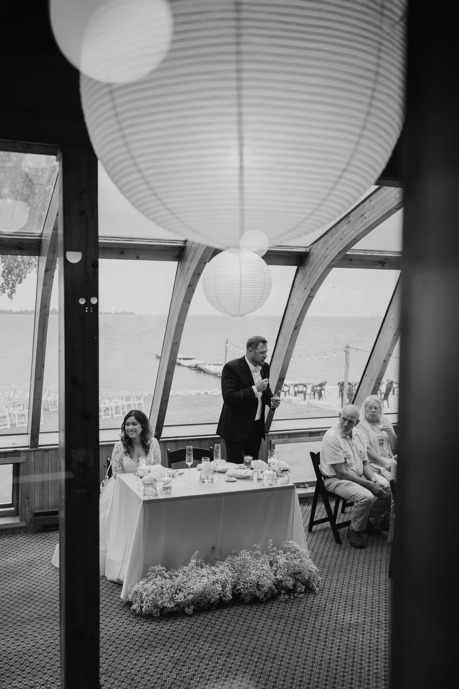 Black and white wedding reception photo with groom giving toast at sweetheart table, paper lanterns, waterfront venue.