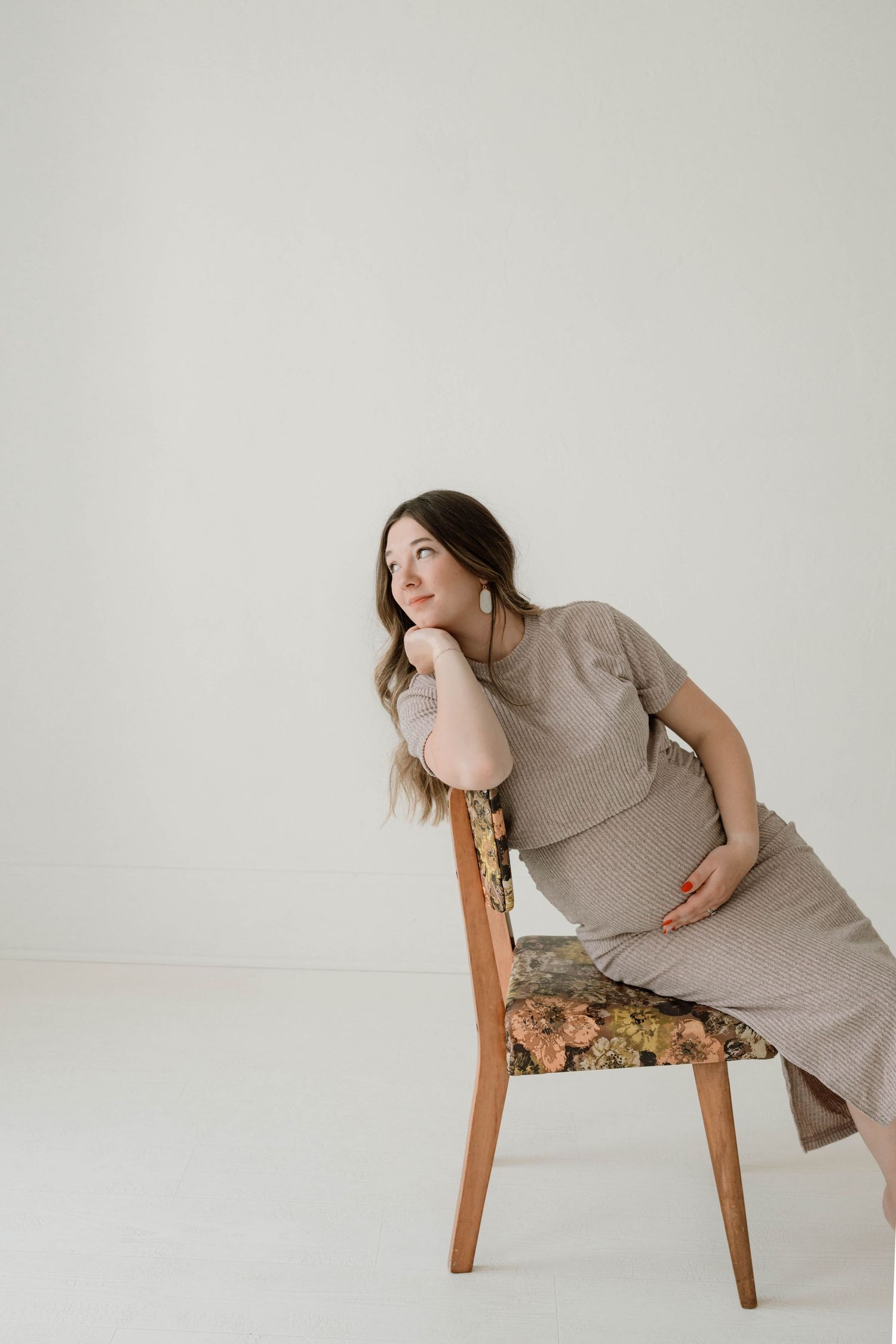 Pregnant woman in beige ribbed dress sits on floral chair, resting chin on hand, gazing upward in bright studio.