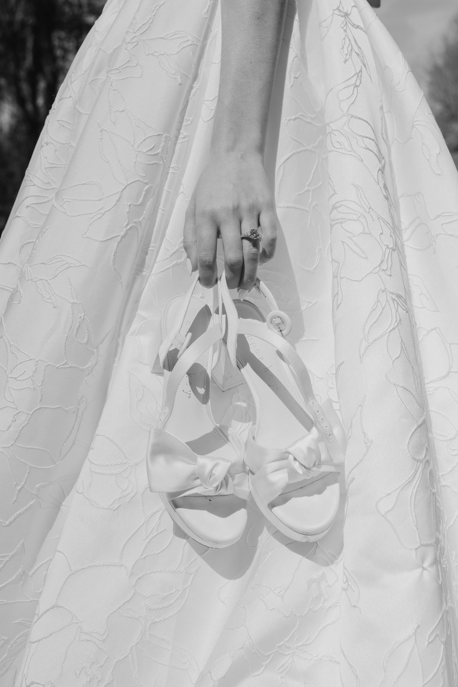 Close-up black and white photo of a bride holding white strappy heels beside her embroidered wedding dress.