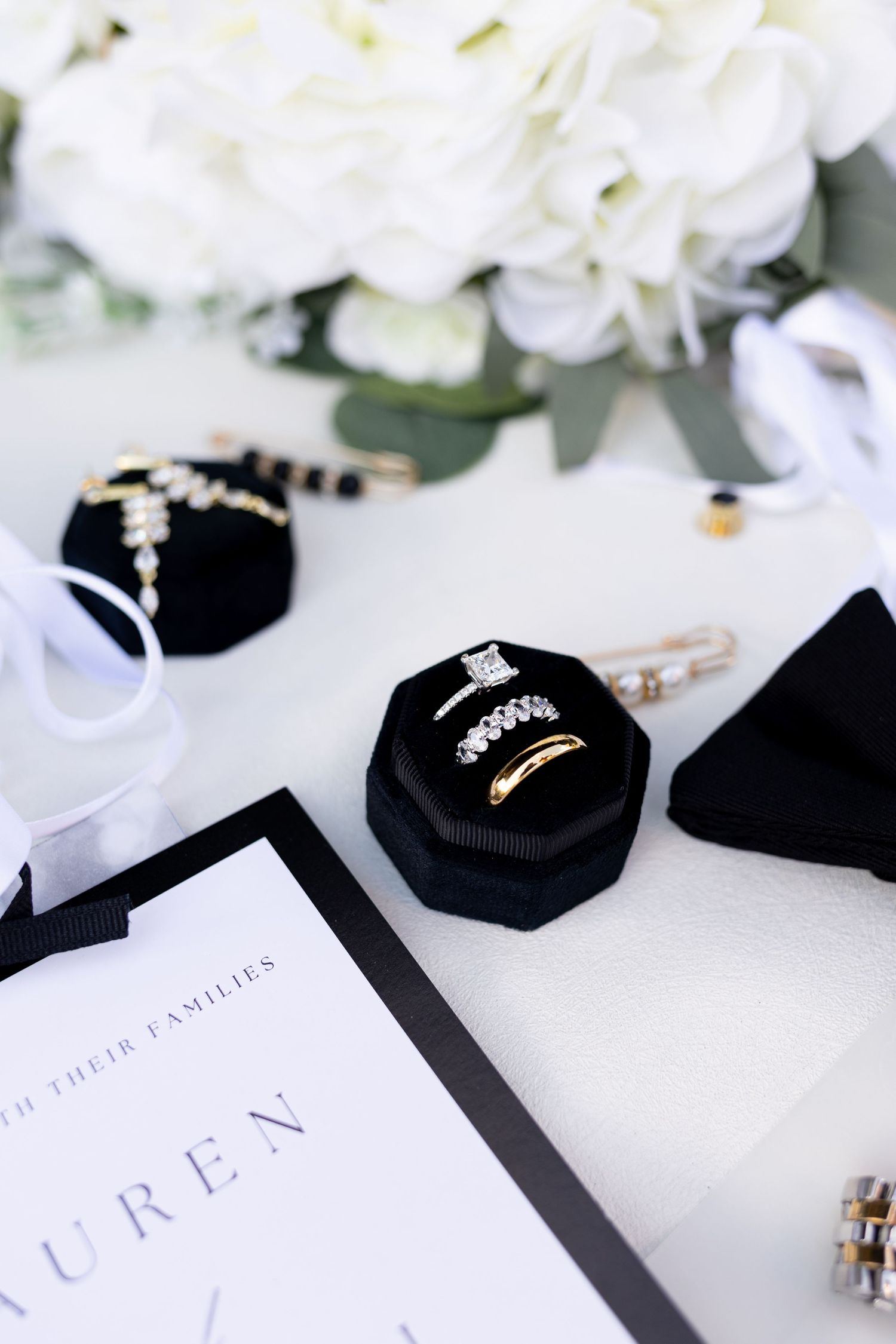 Wedding rings in a black velvet box surrounded by white flowers, jewelry, ribbon, and an invitation.