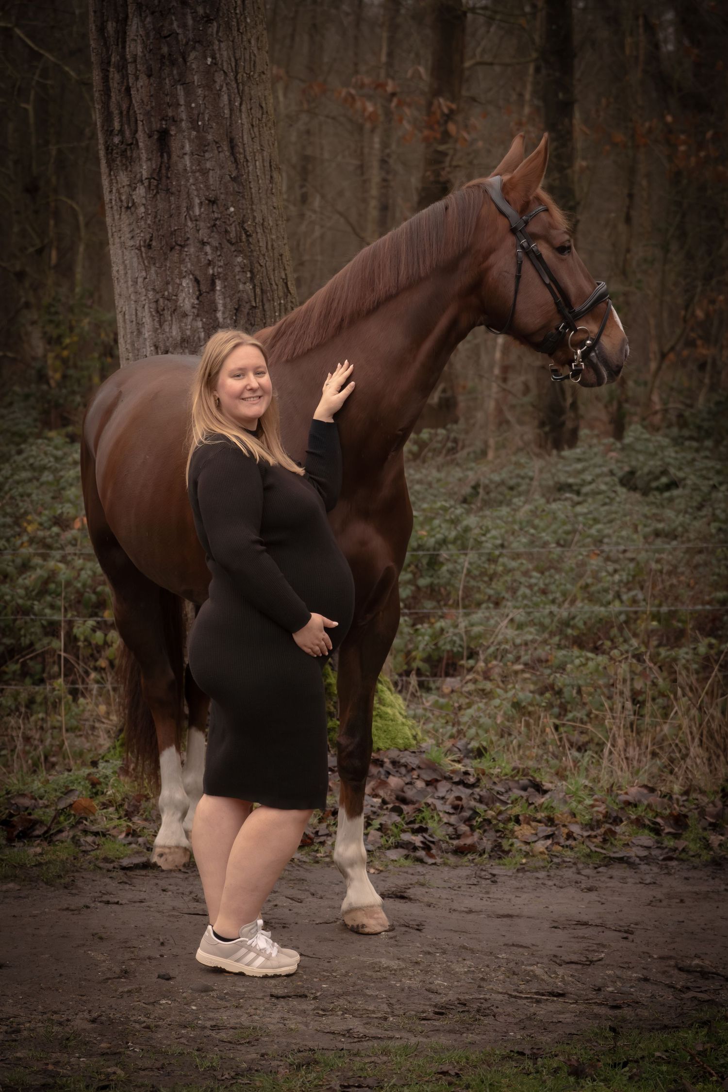 A person in black attire crouches down beside a large brown horse in a wooded setting.