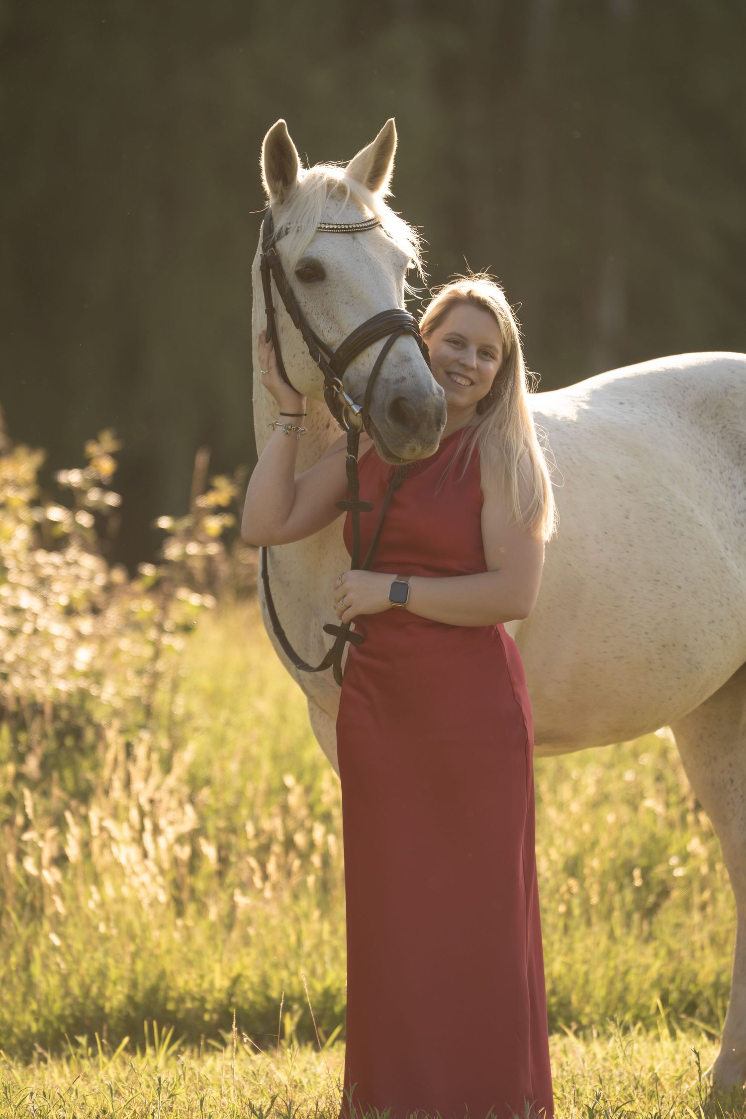 A person in a red dress stands lovingly with a white horse in a sunlit meadow filled with golden grass.