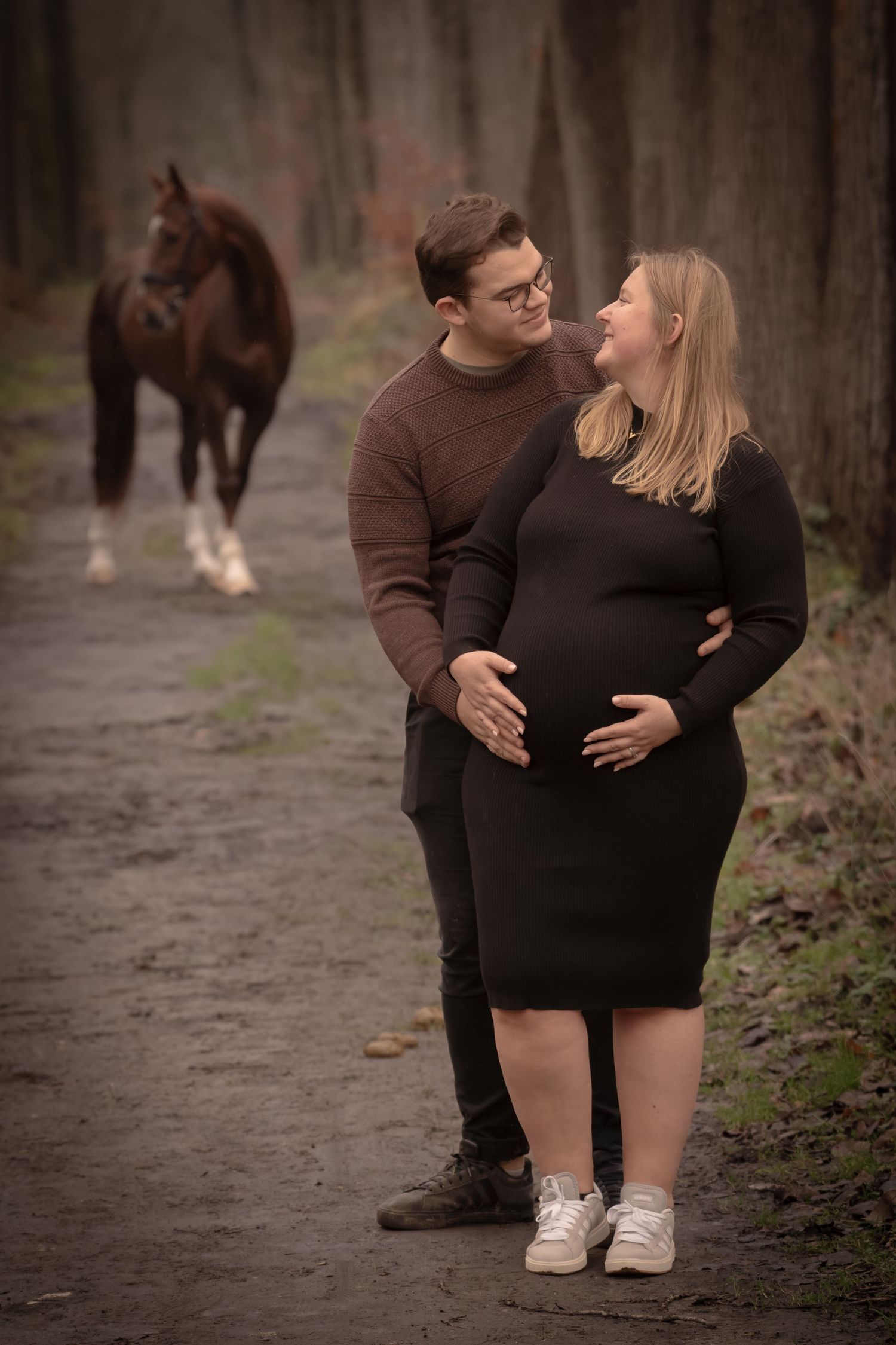 A couple shares intimate moments during a maternity photoshoot on a wooded path with horses in the background.