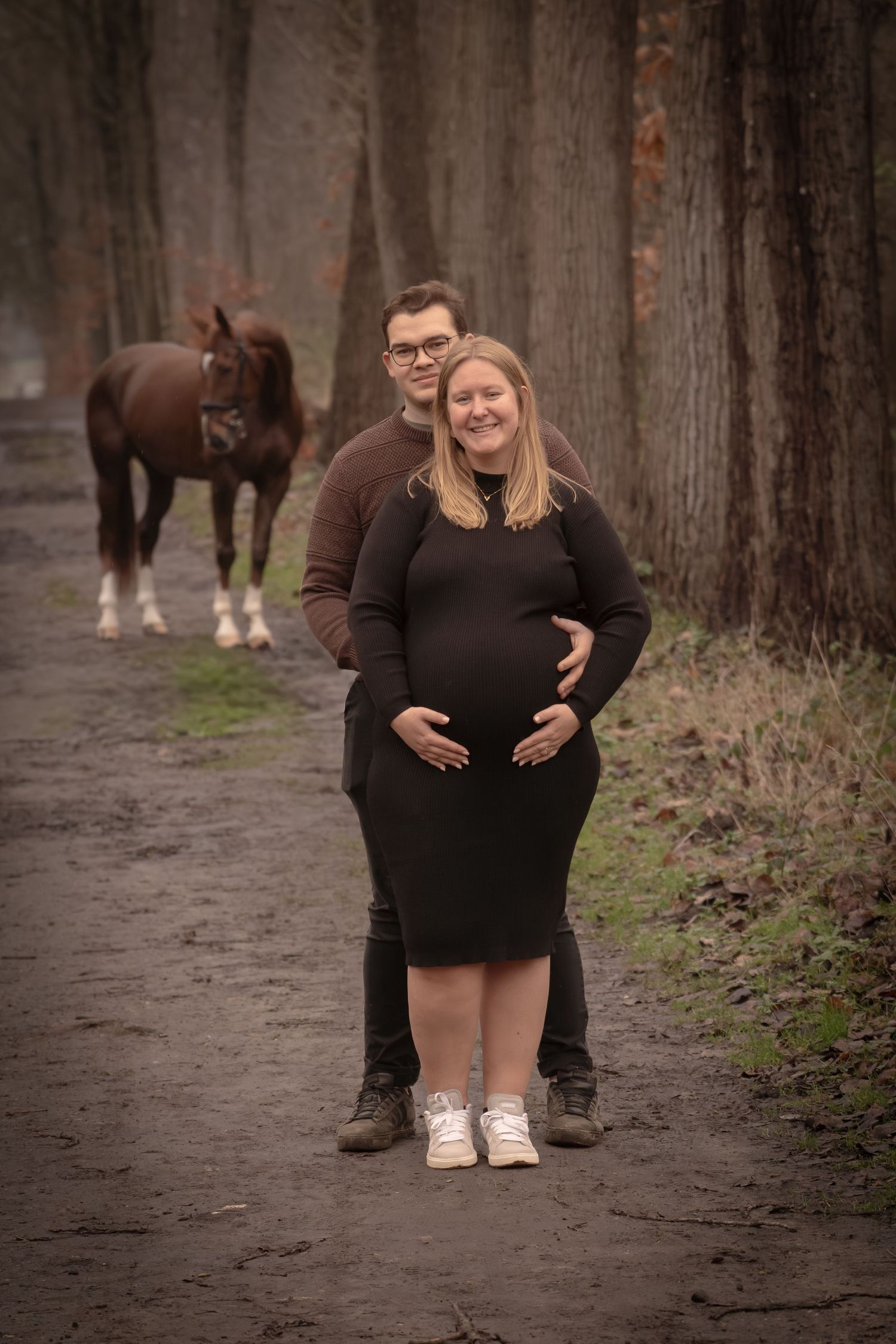 A couple poses together on a wooded path with horses visible in the background.