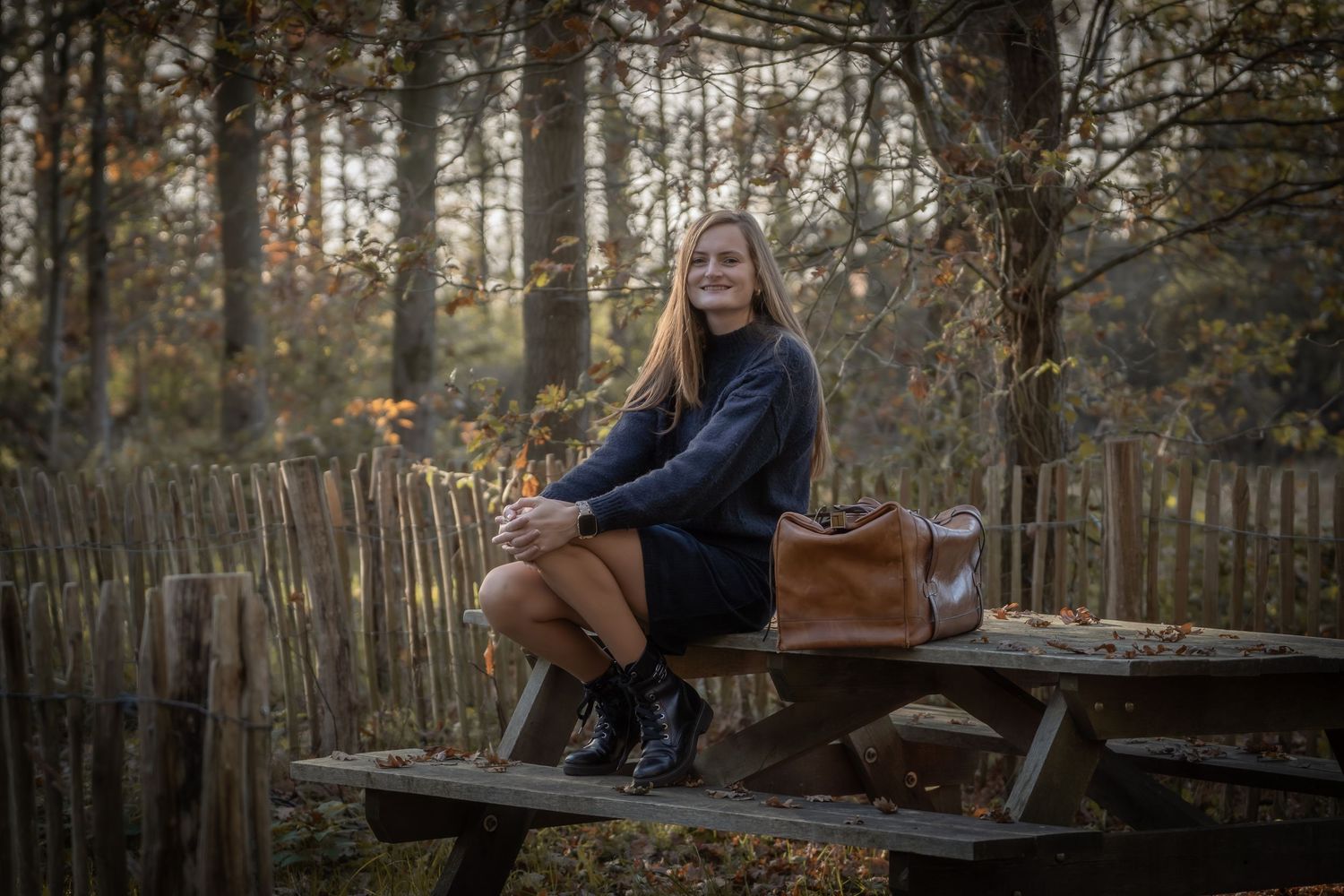 Person sitting on wooden bench in autumn forest surrounded by fallen leaves and trees.