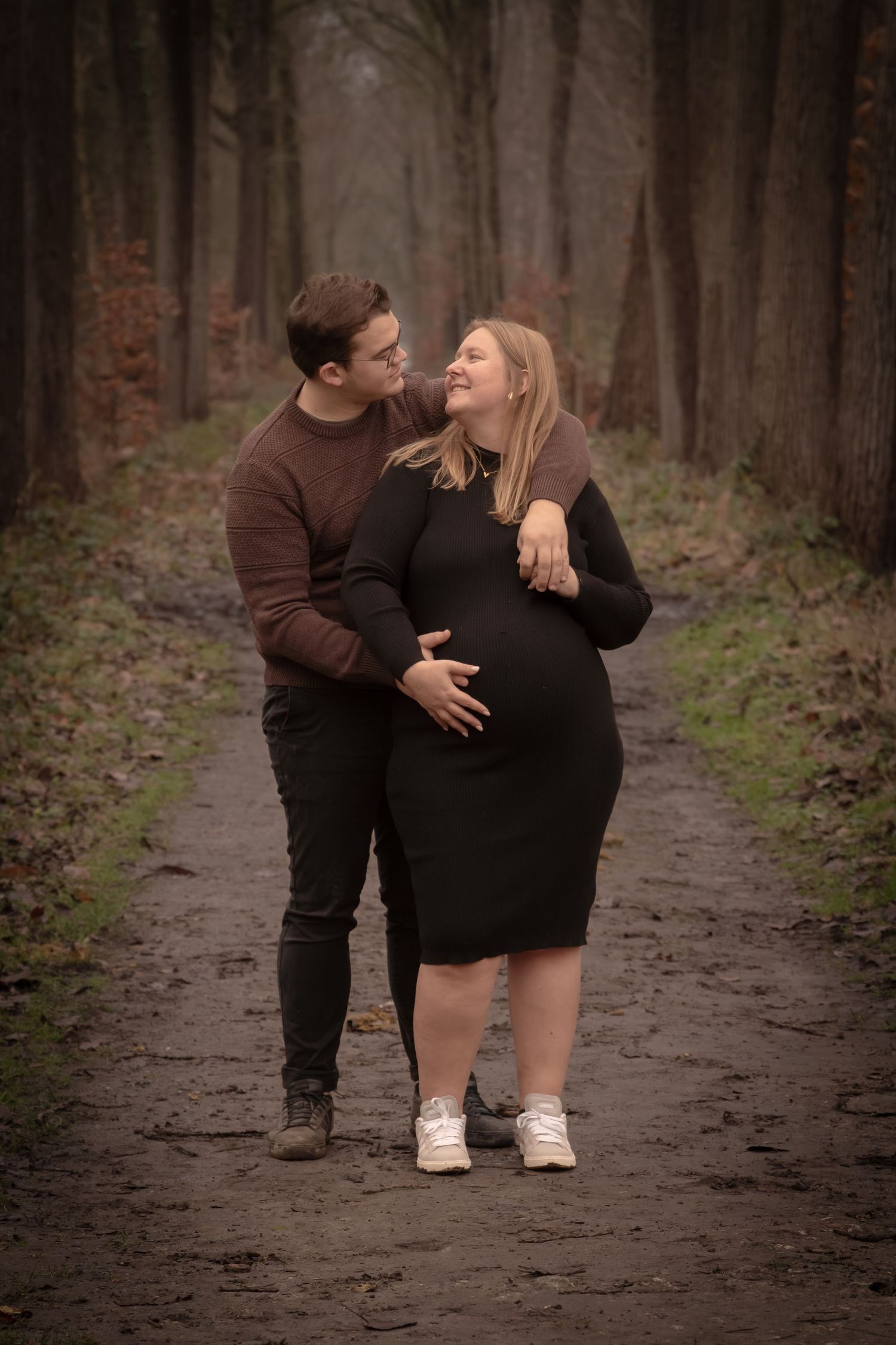 A couple shares an intimate moment on a wooded path surrounded by tall trees in soft lighting.