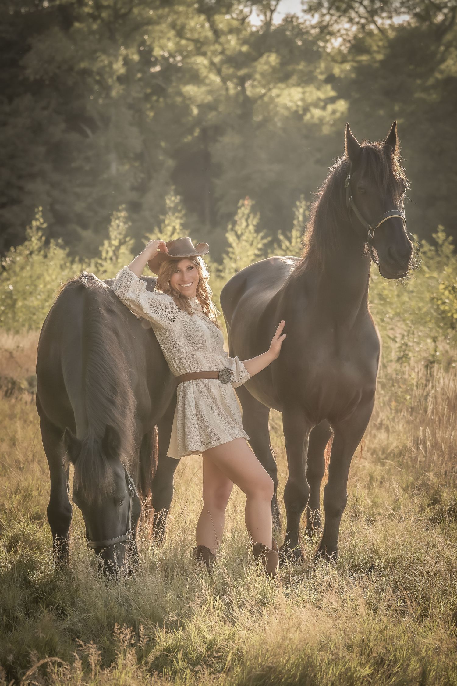 Two majestic black horses graze in a sunlit meadow during golden hour as someone stands between them in a white dress.