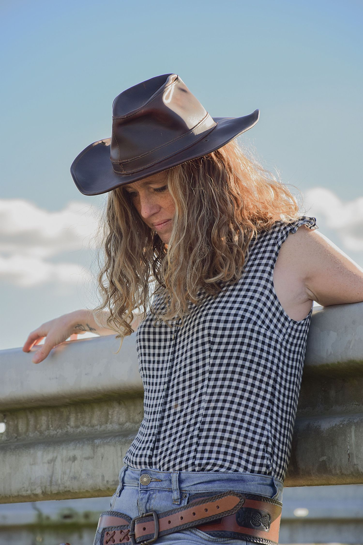 Casual poses against a railing wearing a baseball cap and gingham top.