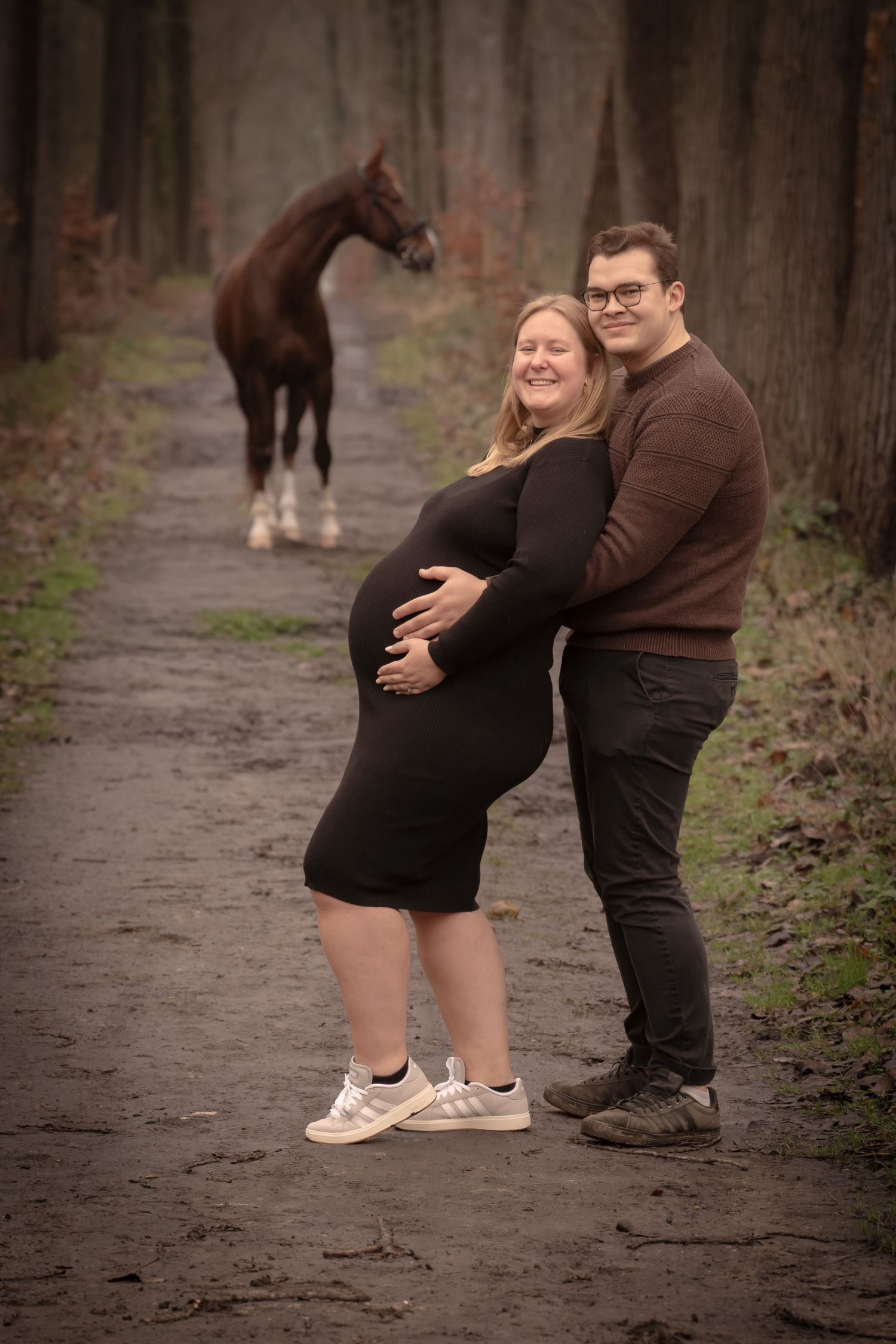 A couple shares a tender moment on a wooded path as they pose for maternity photos in casual attire.