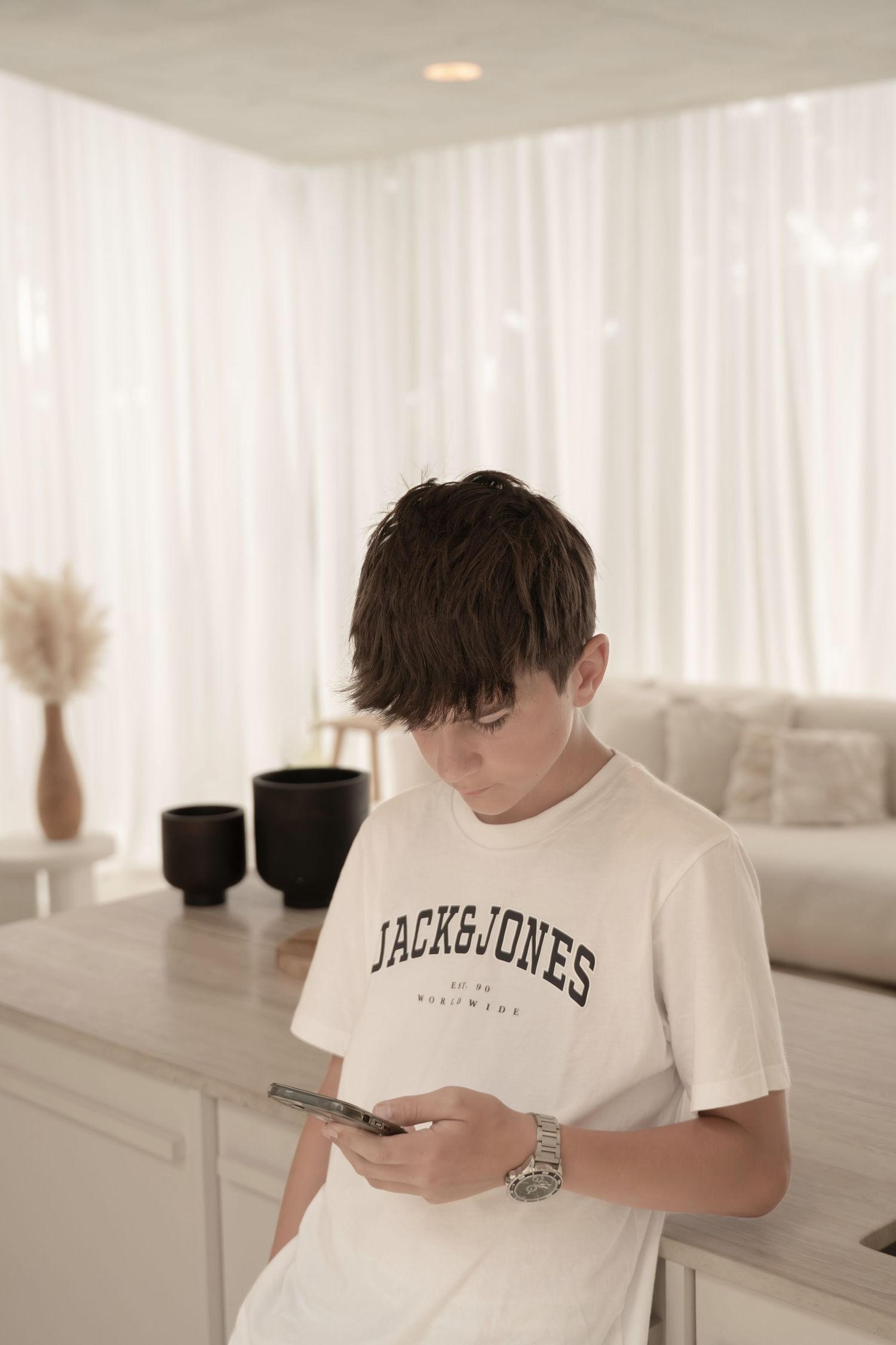 Person in white Jack & Jones t-shirt looking down at mobile phone in minimalist white interior setting.