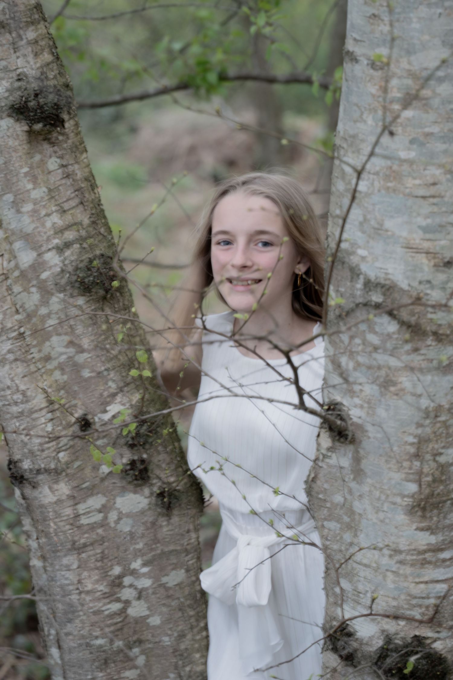 Person in white dress poses between two birch trees in a natural setting.