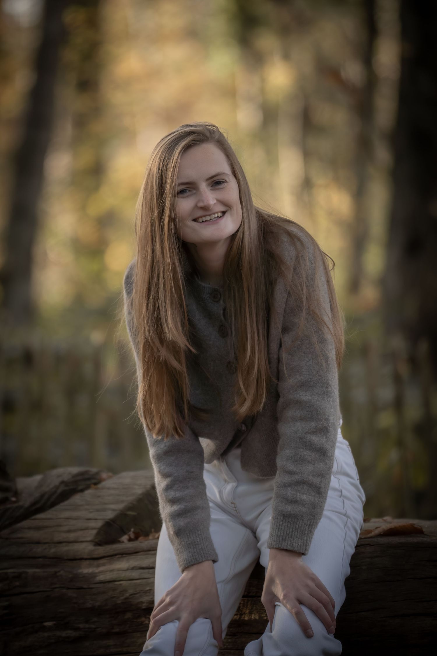 A person in a white sweater and gray cardigan sits on a fallen log in an autumn forest setting.