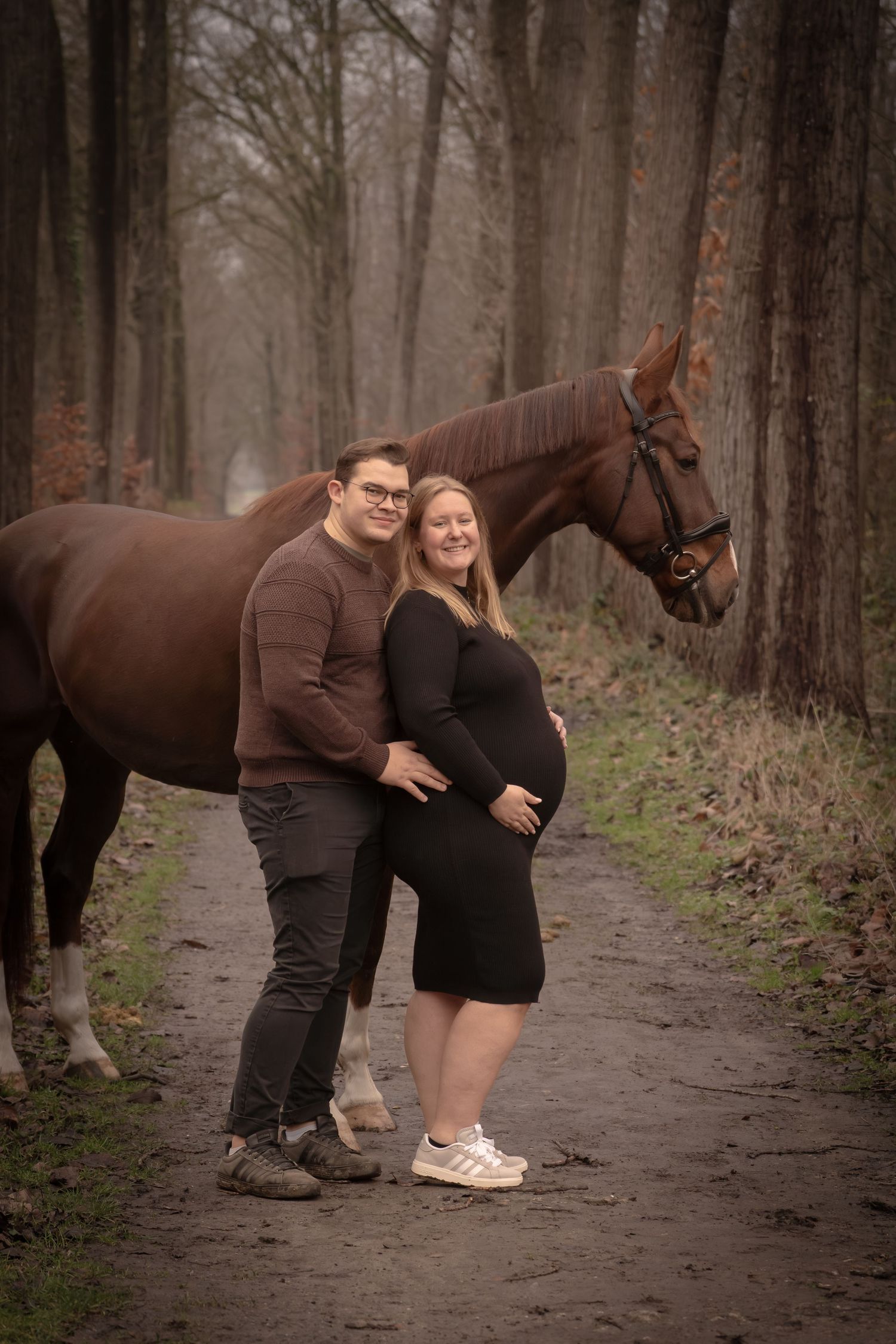 A couple poses together on a misty forest path with their beautiful brown horse in autumn.