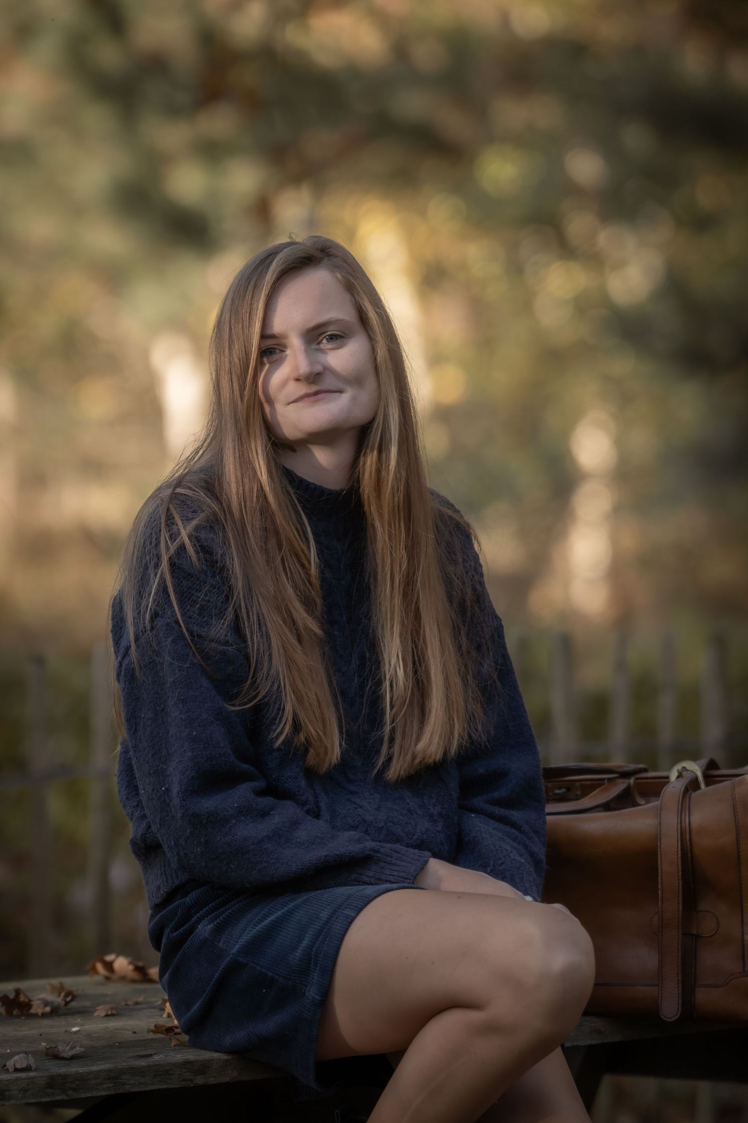 A person in a navy sweater sits casually on a wooden bench outdoors with autumn foliage in the background.