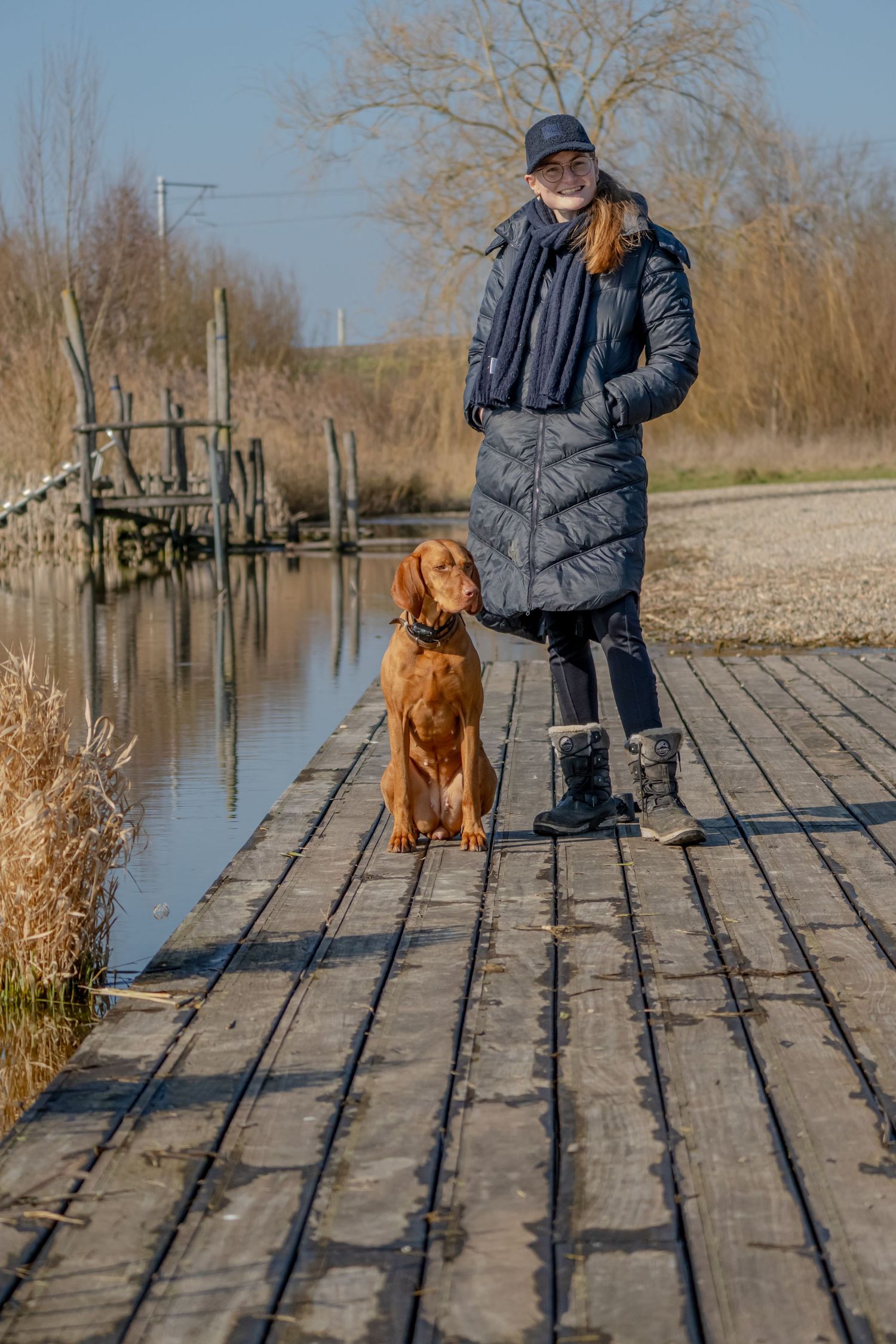 Person in winter coat stands with brown dog on wooden dock by water.