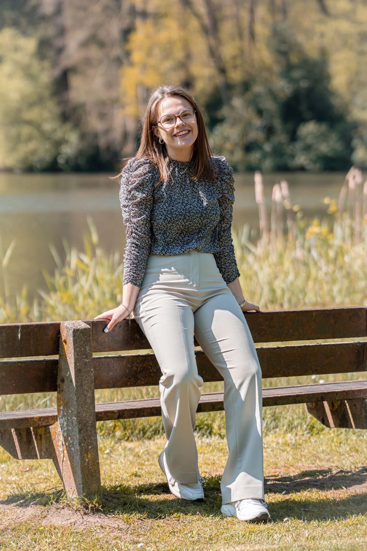 Person in printed top and light pants sits on wooden fence by autumn lake.