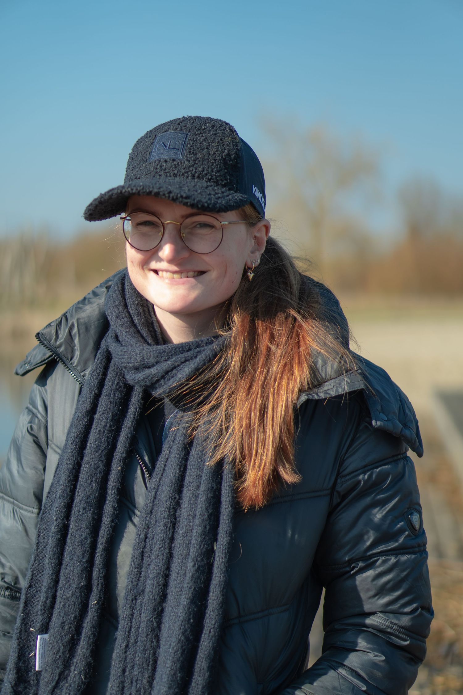 Person wearing winter coat and baseball cap smiles outdoors on cold day.