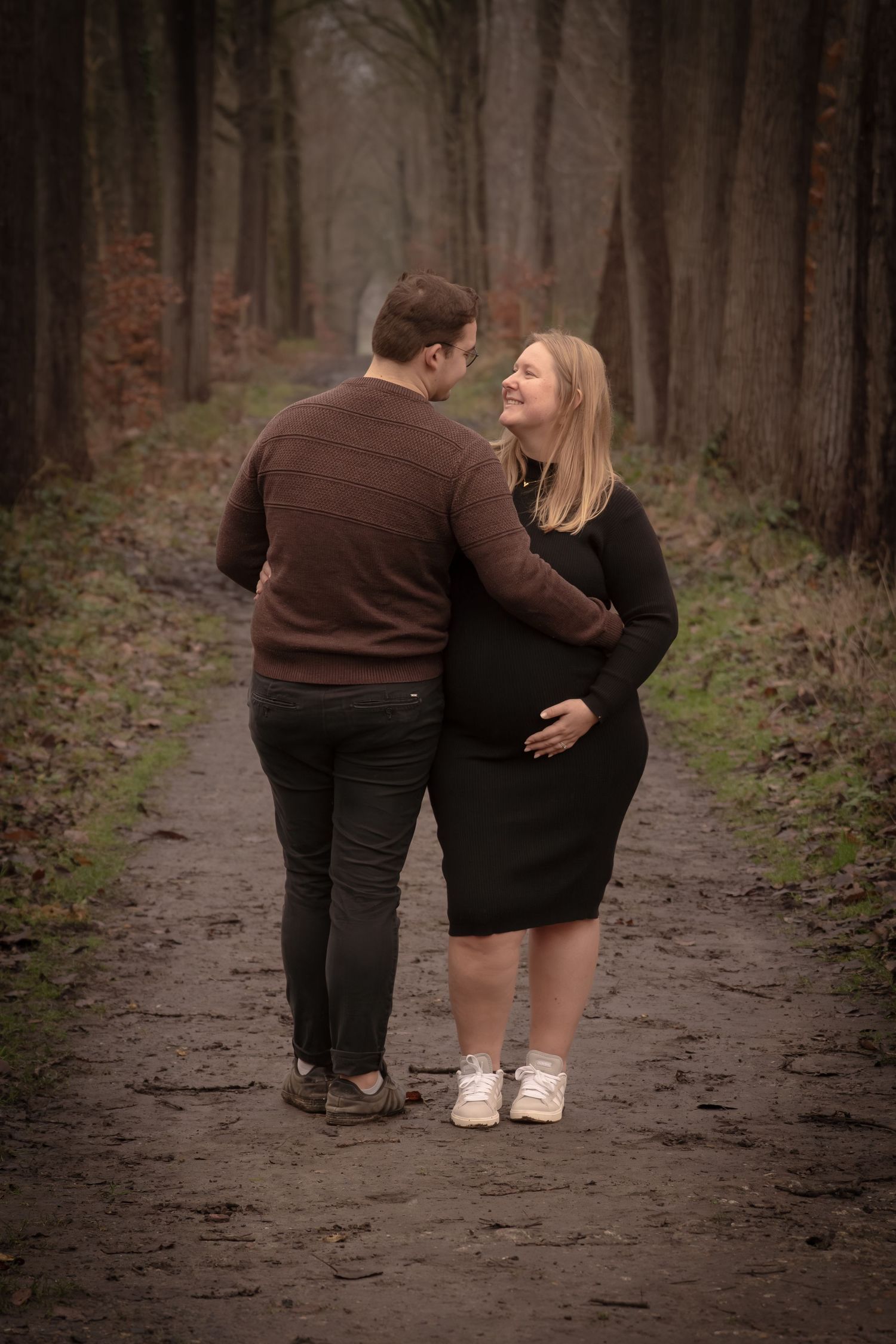 A couple in dark clothing walks together on a forest path during autumn, shown from behind in a romantic moment.