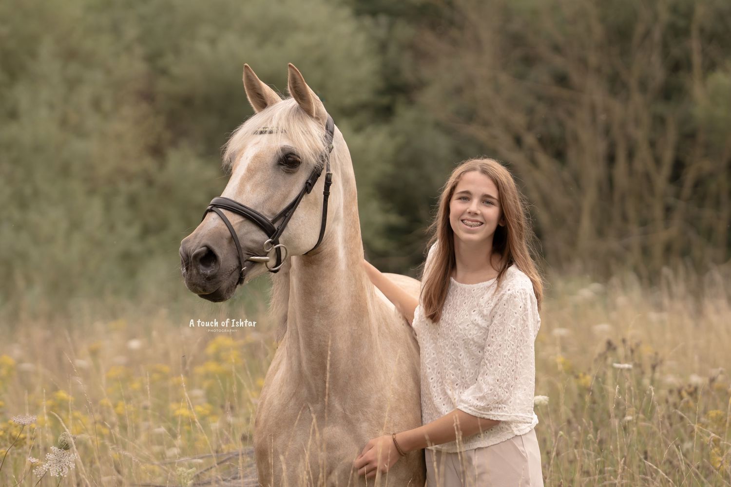 A cream-colored horse and rider stand in a sunlit meadow filled with tall grass and wildflowers.