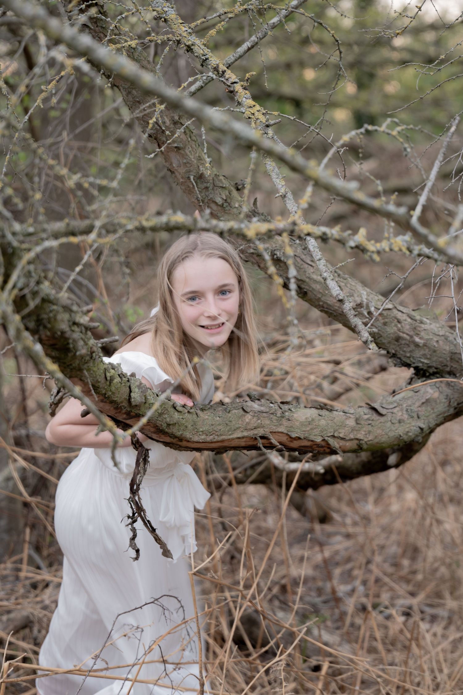 Person in white dress poses among tree branches in a rustic outdoor setting.