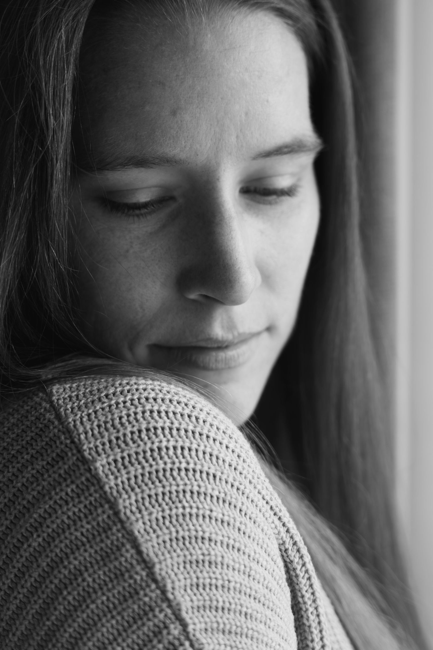 Black and white portrait of person in sweater looking down pensively by window.