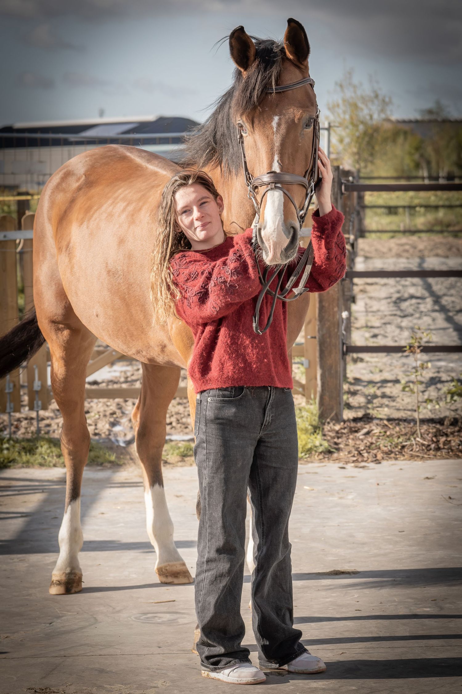 A brown horse stands with its handler wearing a red sweater in a fenced outdoor arena on a cloudy day.