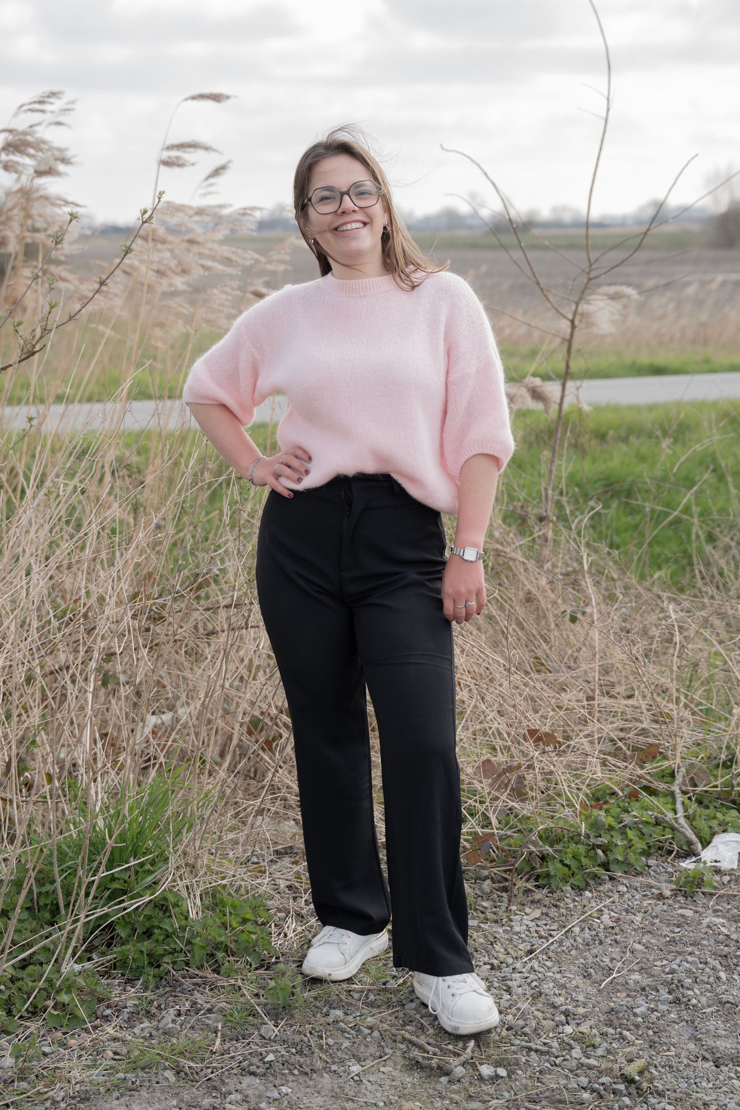 Pink sweater and black pants outfit photographed outdoors among tall grass on a cloudy day.