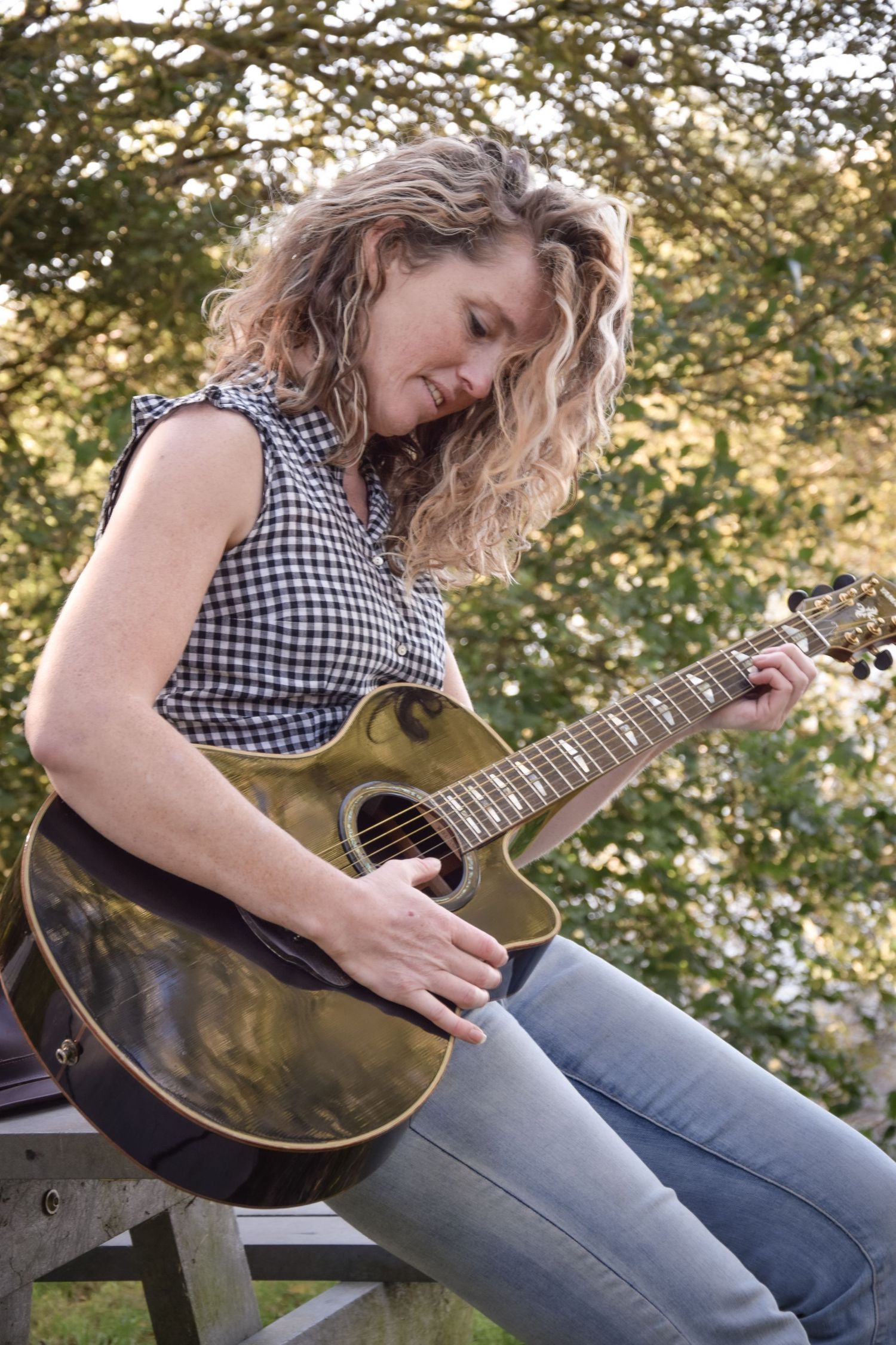 Person playing an acoustic guitar while sitting outdoors wearing a checkered top.