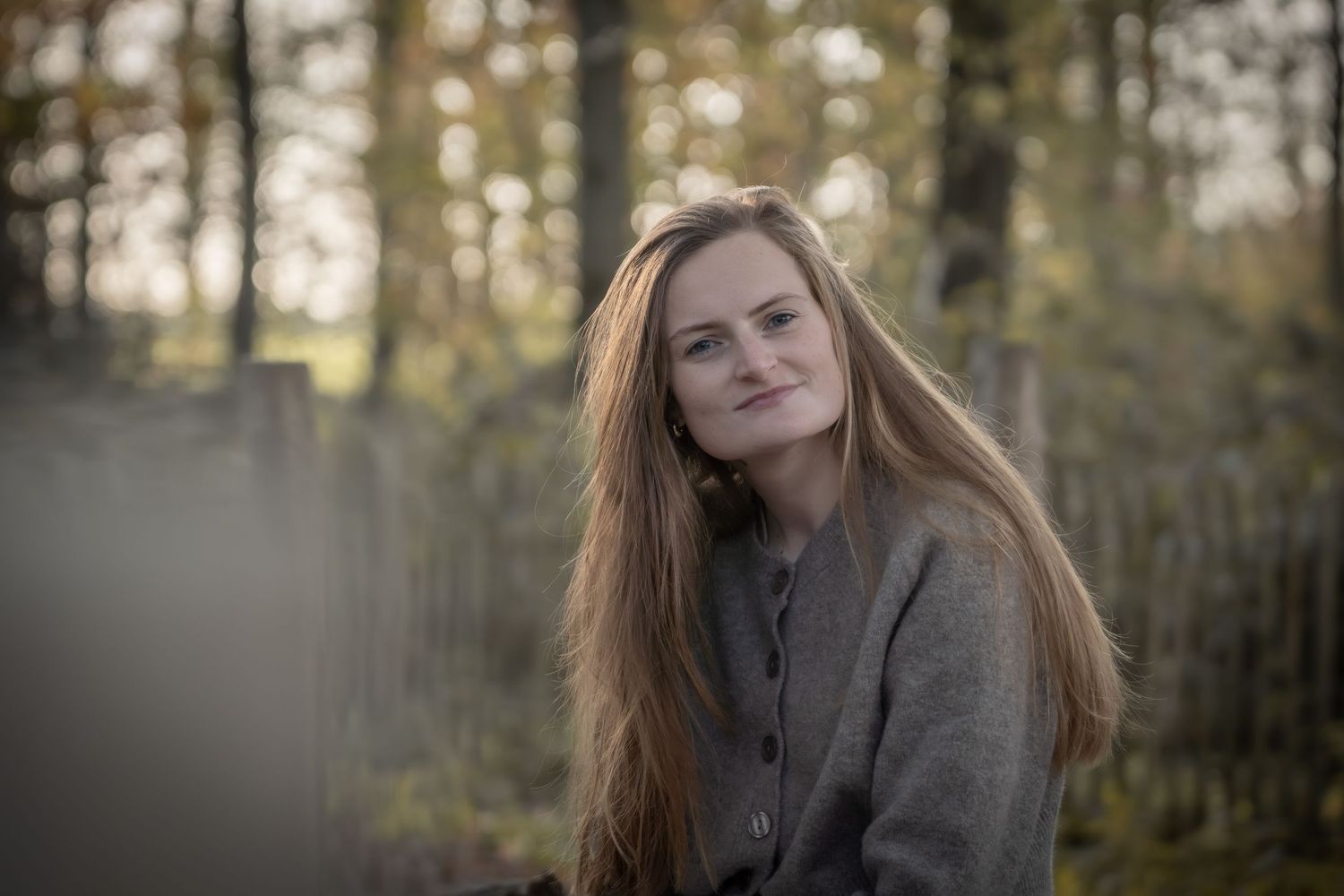 A person with long red hair stands in a forest setting wearing a gray coat during golden hour lighting.