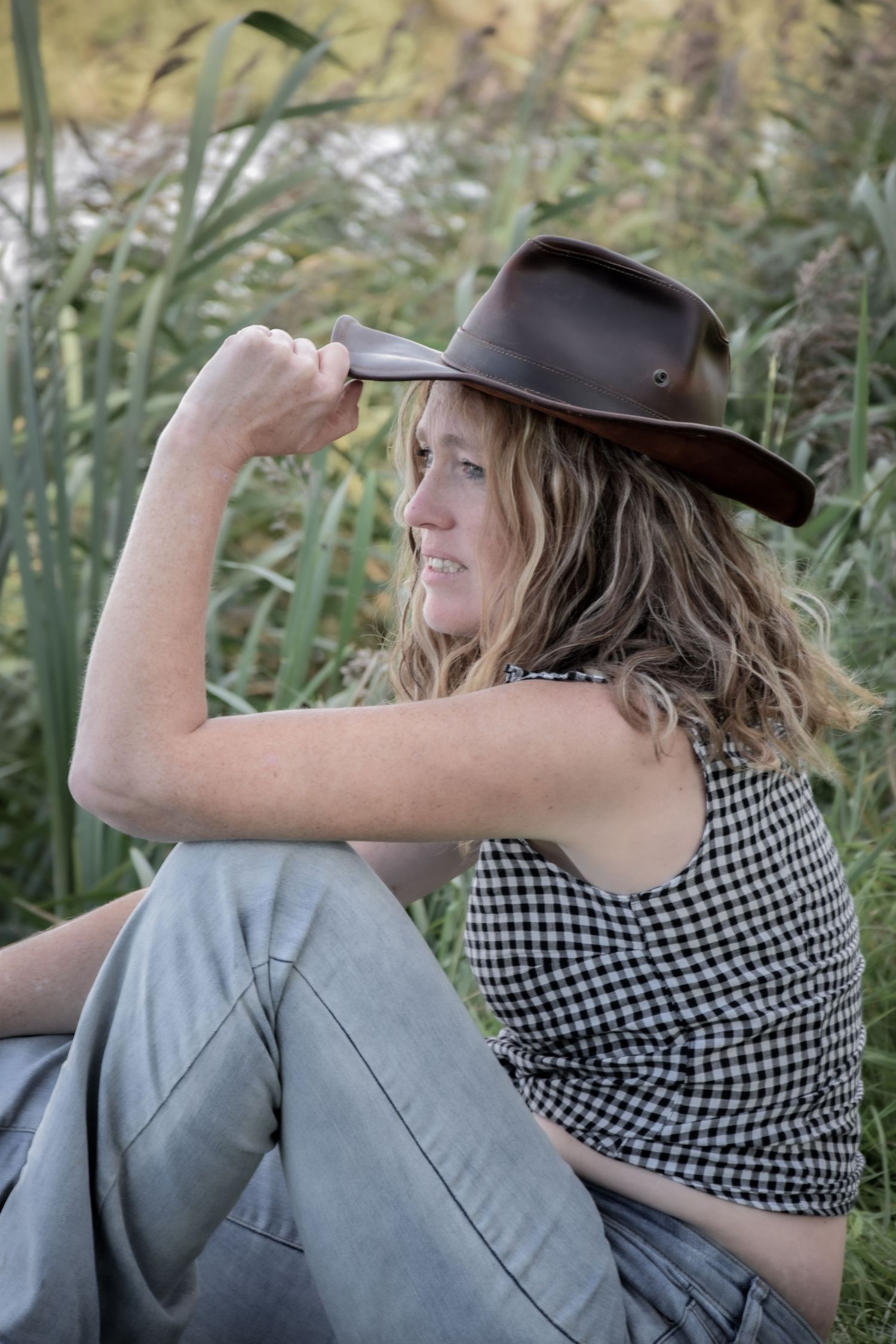 Sitting on a bench in a field wearing a cowboy hat and gingham top.