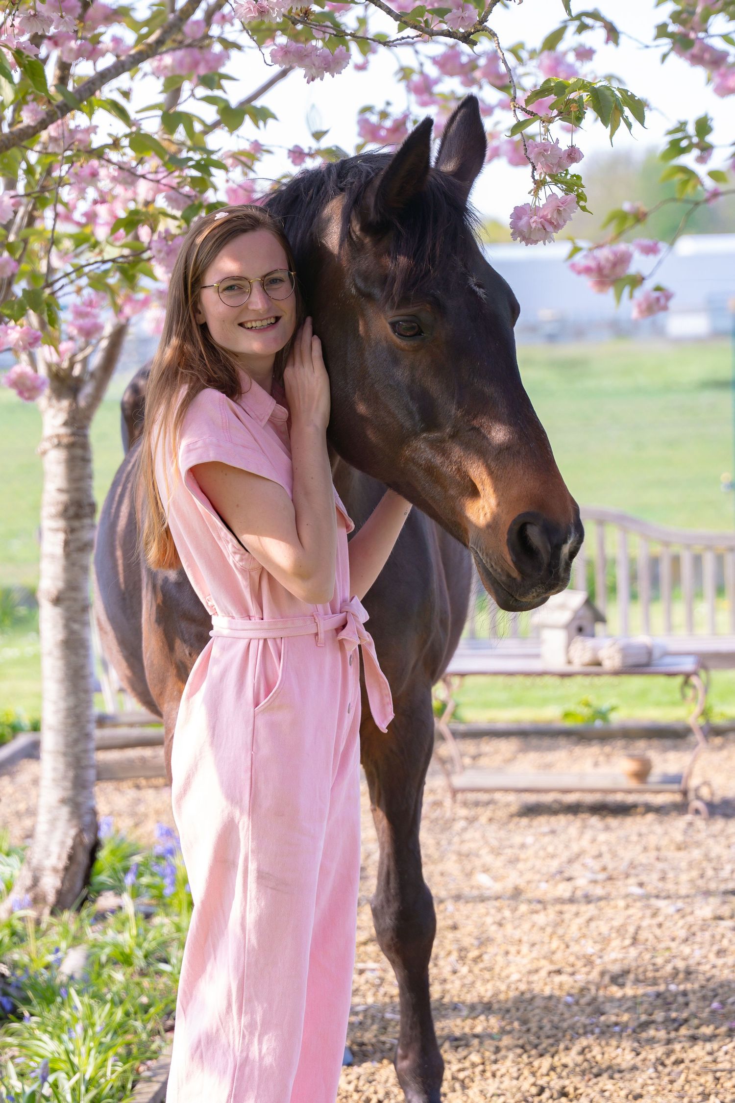 Person in pink jumpsuit poses with dark horse under blooming cherry blossom trees.