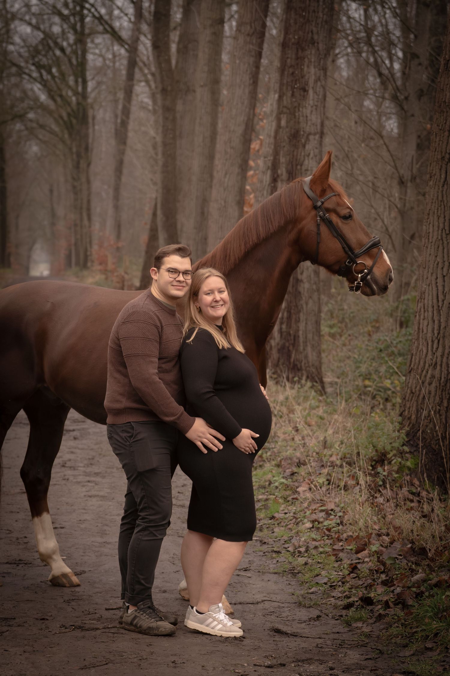 A couple poses with their brown horse on a wooded path during an outdoor photoshoot session.