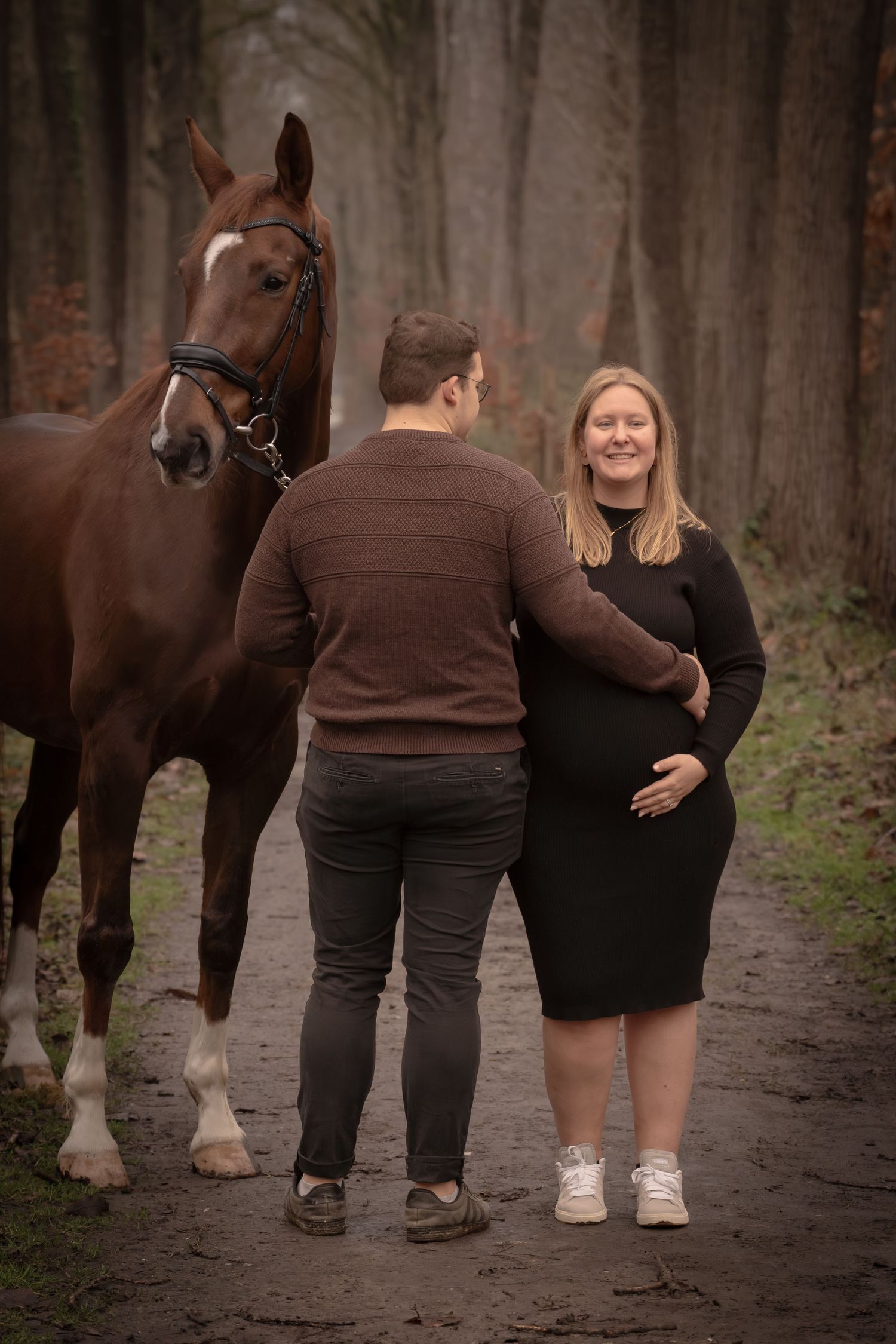 A couple stands together on a forest path with their beautiful bay horse on an autumn day.